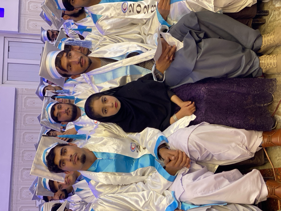 A vertical shot depicts a gathering of people, likely graduates, in white and blue gowns and caps. A young girl in a black hijab sits in the foreground, contrasting with the graduates. The setting appears to be indoors, potentially an auditorium or hall. The graduates are seated closely together, filling the frame, with the focus on the girl in hijab.