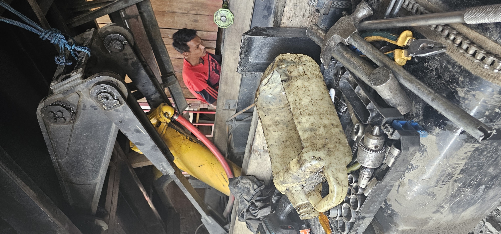In Puncak Jaya, Indonesia, a mechanic, a male with dark hair and skin, is bent over a piece of heavy machinery, his red and black shirt contrasting with the grimy environment. He appears to be working on the machinery, which has a large yellow hydraulic cylinder and a red hose connected to it. Scattered around are various tools, including a dirty plastic container, a toolbox filled with wrenches and sockets, a drill bit, pliers, a hammer, and a chain. The overall scene suggests a workshop or a maintenance area, possibly outdoors, given the wooden planks visible in the background and the accumulation of dust and dirt on the equipment and tools. The lighting is somewhat dim, indicating it might be overcast or the area is under cover.