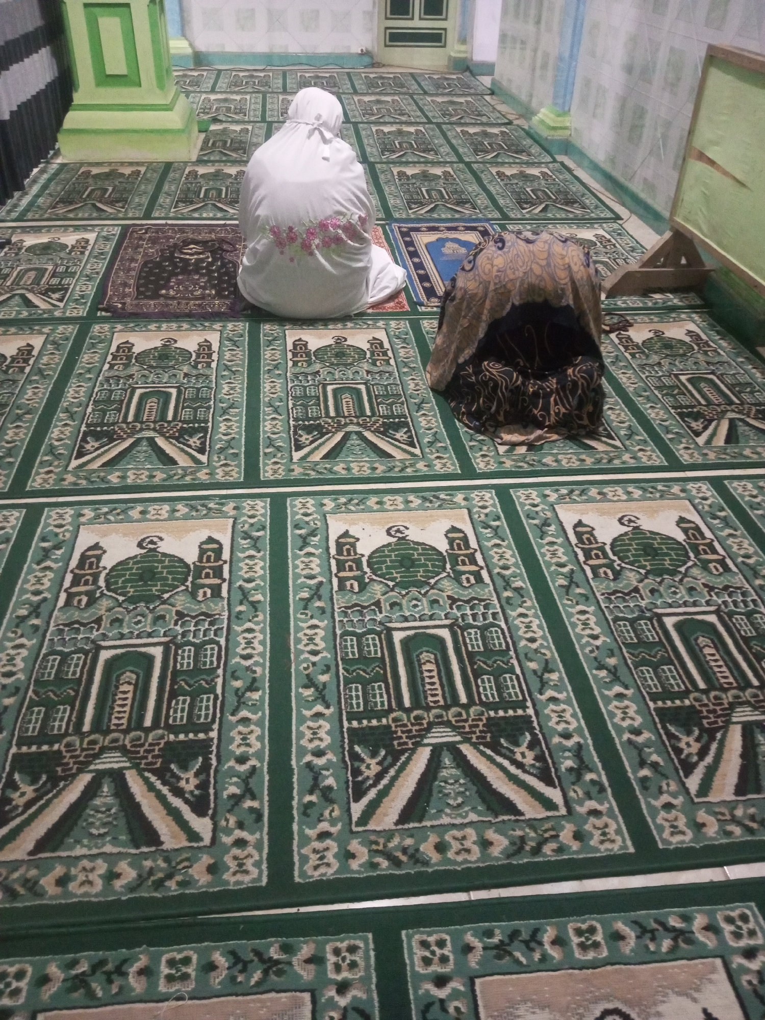 In a mosque in Andongsili, Indonesia, two individuals are engaged in prayer. The carpeted floor is adorned with a repeating pattern of green mosque motifs, with individual prayer rugs placed upon it. One person, clad in a white prayer garment, is positioned in the foreground, facing away from the camera. To their right, another individual is also in prayer, covered by a patterned hijab. The mosque's interior features light-colored walls and a green pillar in the background. The scene is captured from a high-angle perspective.