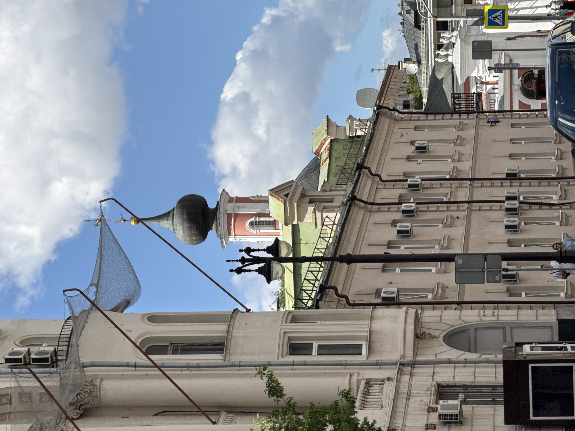 The photograph captures a street scene in Moscow, Russia, on a bright, cloudy day. The perspective is at eye-level, looking slightly upward.
Dominating the left side of the frame is a light-colored building with architectural details such as a decorative cornice and arched windows. Two outdoor air conditioning units are mounted on its facade. A metal net, supported by red poles, is stretched out from this building towards the right.
In the mid-ground, a multi-story building with a pale pink facade runs horizontally across the image. This building also features numerous air conditioning units and regularly spaced windows. Above this building, a green-roofed structure is partially visible.
Further to the right, a prominent church spire with a dark onion dome topped by a cross rises against the blue sky. The spire has a red and white section at its base.
In the foreground, a black street lamp with two globes stands on the right side of the street. A pedestrian crossing sign, a blue square with a white pedestrian symbol, is visible on a pole behind the street lamp.
Two individuals, one male in a white t-shirt and one female in a blue shirt, are standing near the pedestrian sign. Further in the distance, other buildings and structures, including a white decorative fence and gate, are visible. A dark-colored car is partially visible in the lower right corner, suggesting it is on the street. Lush green foliage from a tree is present on the far left. The sky is filled with fluffy white clouds.
