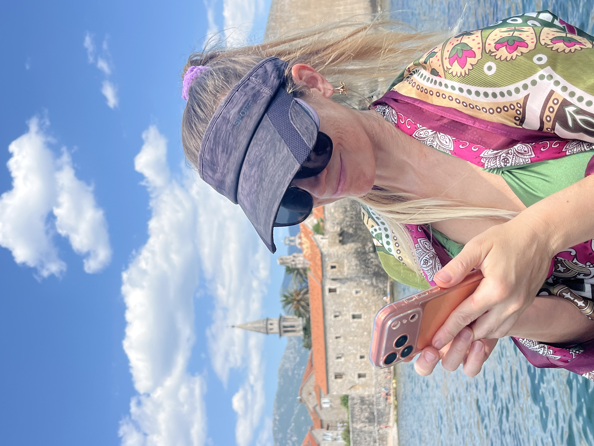 A woman, wearing a visor and sunglasses, holds a rose-gold smartphone in her hands, her fingers are wrapped around the device. She is looking down at the screen. Behind her, a scenic view unfolds, featuring a coastal town with stone buildings and a prominent church steeple. The sky is a bright blue with scattered white clouds. The water in the foreground appears calm and reflective. The woman is dressed in a vibrant, patterned scarf or cover-up worn over a green bikini top. Her blonde hair is visible and partially pulled back by a purple scrunchie. The scene suggests a sunny day, likely during daytime, in a Mediterranean or coastal location. The text "SPORT" is visible on the visor. The background scenery, with its distinctive architecture and seaside setting, is consistent with Budva, Montenegro. The woman's focus on her phone indicates she is likely engaged in communication or browsing.