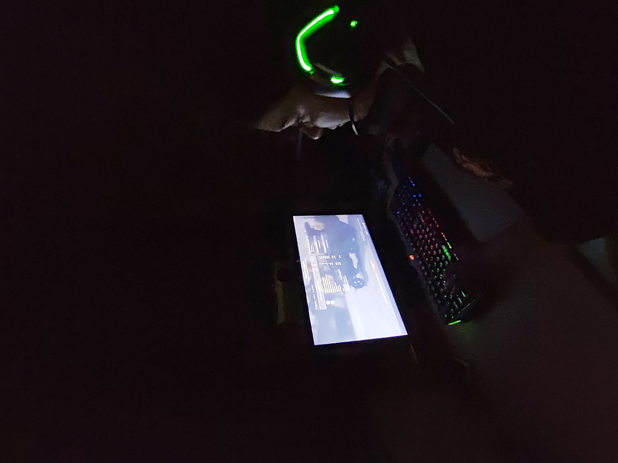 A dimly lit scene shows a person wearing a headset with green glowing lights, looking down at a tablet screen. To the right of the tablet is a gaming keyboard with multicolored backlighting. The environment is dark, suggesting an indoor setting, possibly at night. The location is not discernible from the image.