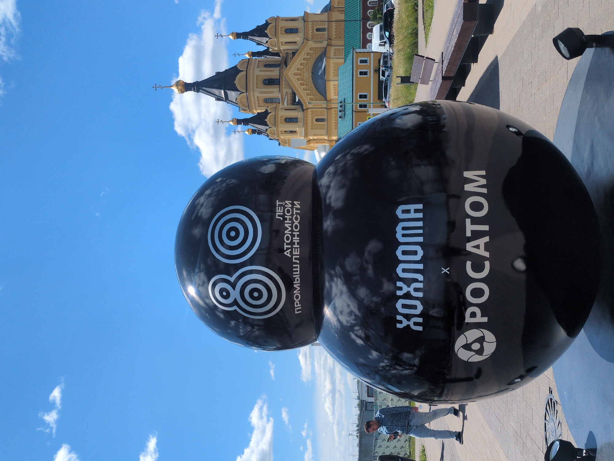 This is a photograph taken at the Nizhny Novgorod Kremlin, Russia. In the foreground, a large, black, spherical sculpture is prominently displayed. The sculpture has two rounded sections, one atop the other. The upper section features a large stylized "80" in silver, with smaller concentric circles within the larger circles of the "8". Below this, text in silver reads "ЛЕТ АТОМНОЙ ПРОМЫШЛЕННОСТИ" (Years of the Atomic Industry). The lower section of the sculpture displays the word "ХОХЛОМА" in large silver Cyrillic letters, followed by a stylized "x" and then "POCATOM" in silver Cyrillic letters, with the Rosatom logo. To the left of the sculpture, a man wearing a blue plaid shirt and light-colored jeans is walking while looking at his phone. In the background, the Alexander Nevsky Cathedral, a prominent landmark of Nizhny Novgorod, stands with its golden domes and yellow facade. The sky is bright blue with scattered white clouds. The ground in front of the sculpture is paved with light-colored rectangular tiles. Benches are visible to the right of the sculpture.
