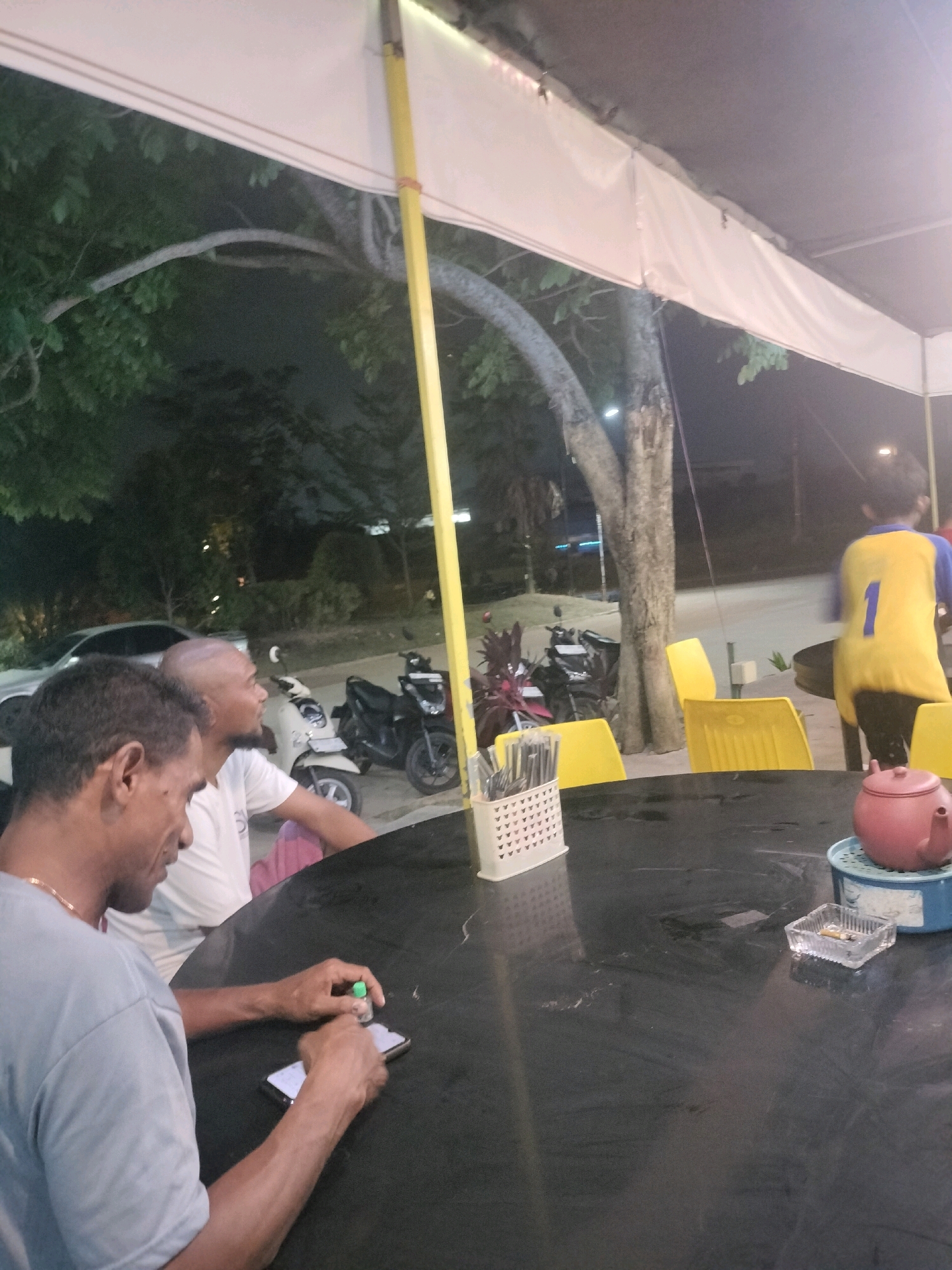 The image depicts an outdoor seating area at night, likely a street-side cafe or warung, in Batam, Indonesia. In the foreground, a man with short, dark hair is seated at a black, round table. He is wearing a grey t-shirt and appears to be interacting with his smartphone, which is on the table. His hands are visible, one holding a small green-capped object, possibly a small bottle of liquid or medication, near the phone. To his left and slightly behind him, another man with a shaved head is seated, wearing a white t-shirt.
The table also holds a white plastic utensil holder filled with cutlery, an ashtray with cigarette butts, and a pink teapot with a blue lid. Yellow plastic chairs are visible in the background. Beyond the table, a few motorcycles are parked. In the middle ground, a tree with textured bark is prominent, and further back, trees and foliage are visible, suggesting a somewhat vegetated environment within the city. The overall lighting suggests it is nighttime, with some artificial lights illuminating the scene. A young person wearing a yellow and blue shirt with the number "1" on the back is walking away from the table in the far right background. The presence of motorcycles, the style of the seating area, and the surrounding vegetation are consistent with common street-side establishments found in Batam.