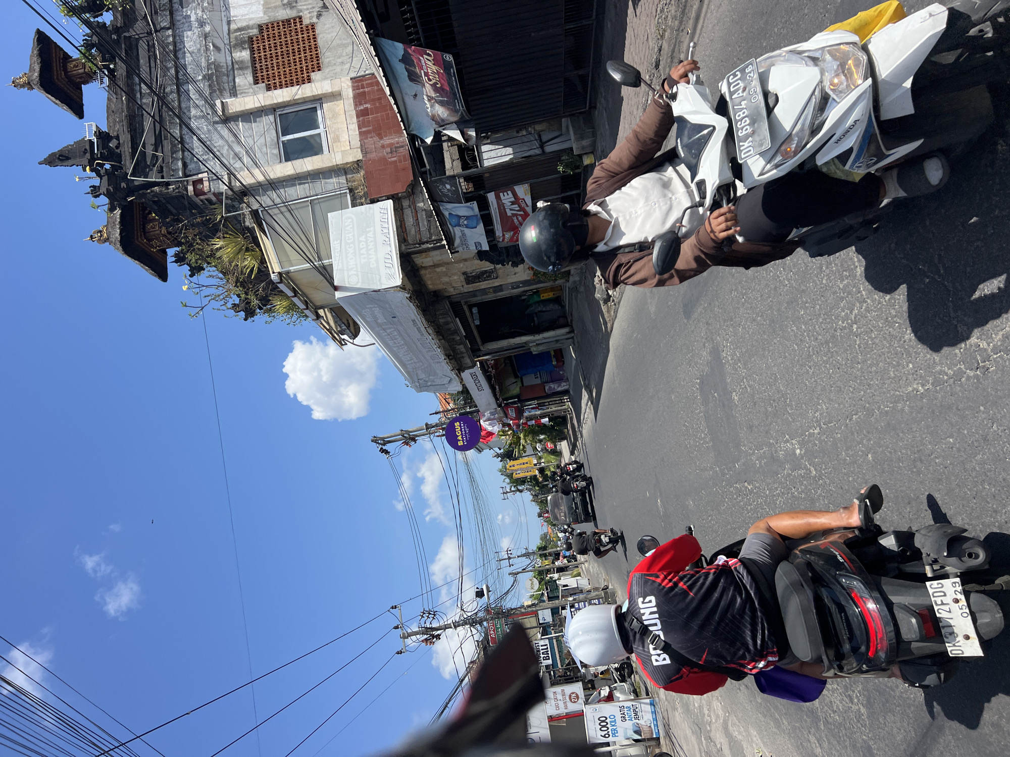 This is a first-person perspective, street-level photograph taken from a motorcycle in Denpasar, Indonesia. The image captures a sunny day with a clear blue sky and scattered white clouds. The foreground features two motorcycles with riders, seen from behind and the side, respectively. The rider in the immediate foreground is wearing a red and black shirt with the word "BOUNG" on the back, grey shorts, and a white helmet. They are on a black scooter with a purple bag hanging off the back. The second motorcycle, to the right and slightly ahead, is white and ridden by a person wearing a brown jacket over a white shirt and black pants, with a black helmet. The street is paved and appears to be a commercial area with buildings lining both sides. These buildings are a mix of styles, some with traditional Balinese architecture, including ornate rooftops and decorative elements, while others are more modern. Various shops and signs are visible, including "APOTHEK", "STATIONERY", and advertisements in Indonesian. Power lines and utility poles are prominent, strung across the street. Other vehicles, including cars and motorcycles, are also present on the road, indicating moderate traffic. The overall scene depicts a typical urban street in Denpasar, Indonesia, characterized by a blend of cultural architecture and everyday commerce.