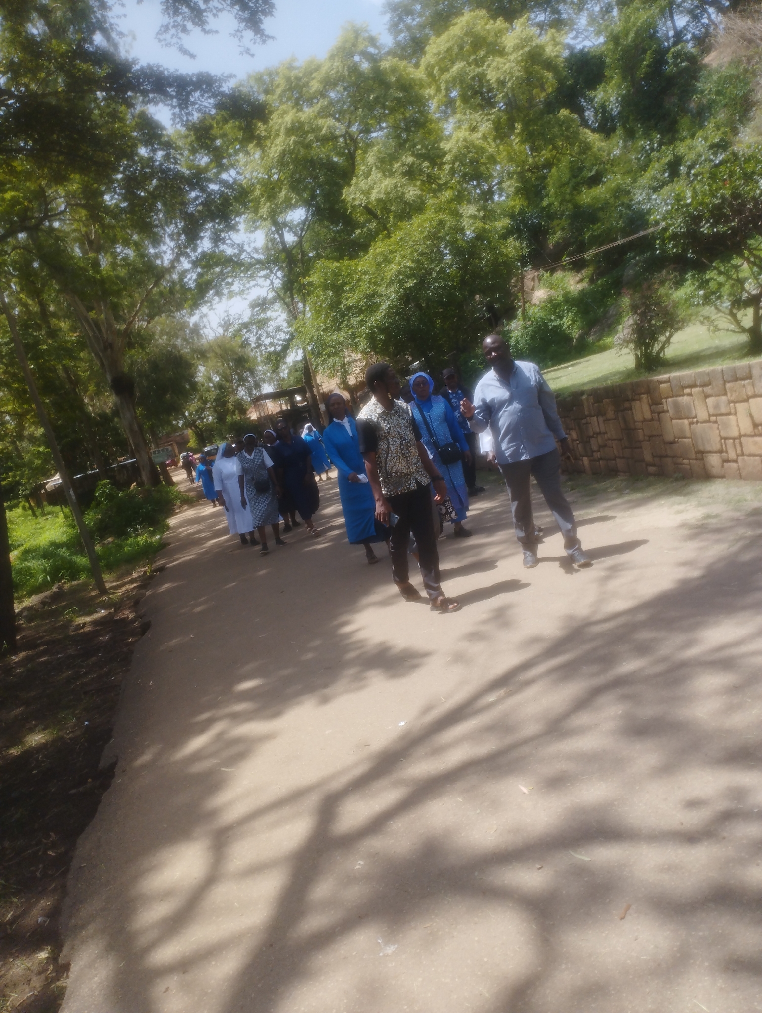 A group of people, predominantly women in blue and white religious habits, are walking on a dirt road lined with trees and a stone wall. The scene appears to be outdoors on a sunny day. The road is dusty and has shadows from the surrounding trees cast across it. The women are walking in a procession, with some in the foreground and others further back. A man in a blue shirt and dark pants is walking separately from the group, facing forward. The environment suggests a rural or semi-rural setting within a city, possibly a church or monastery grounds. The image quality is fair, with some blurriness. It is not possible to determine the specific location within Jos, Nigeria, from the visual information provided.