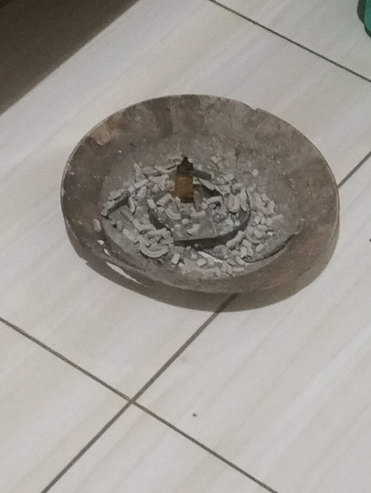 The image shows an ash-filled ashtray on a tiled floor. The ashtray is round and made of a rough, dark material, possibly wood or stone. It contains a significant amount of gray ash and what appear to be remnants of a burnt-out cigarette or incense stick in the center. The floor is tiled with square, light-colored tiles, with dark grout lines forming a grid pattern. A shadow is cast on the floor from an unseen object to the left of the frame. The overall scene is dimly lit. The location is noted as Klecorejo, Indonesia.