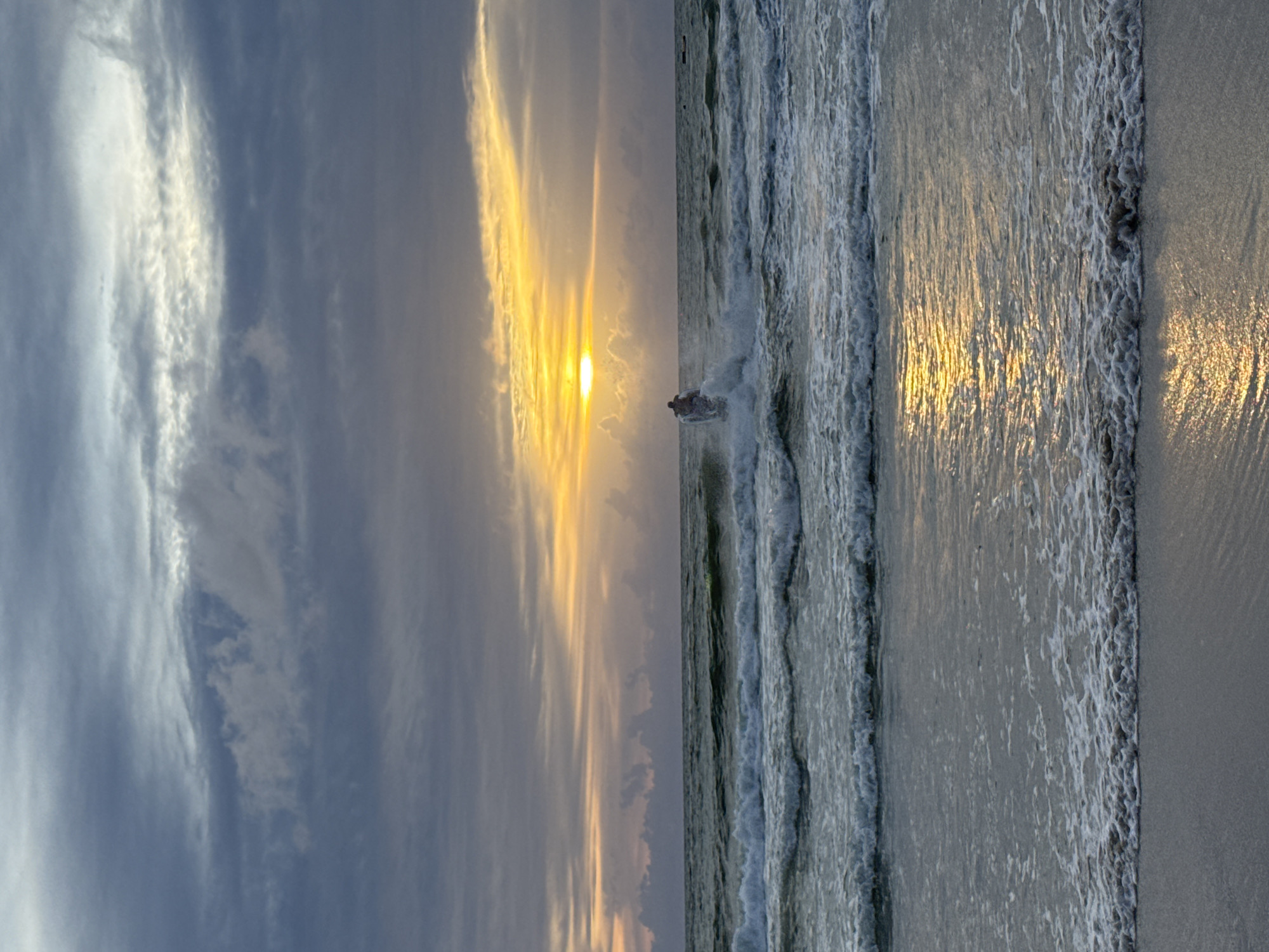 The image captures a serene ocean scene at what appears to be sunset in Patong, Thailand. The sky is filled with dramatic, cloudy formations, transitioning from deep grays to softer blues and yellows near the horizon where the sun is setting. The sun itself is a bright, glowing orb, its light reflecting intensely on the water's surface. The ocean is moderately rough, with visible waves rolling towards the shore, creating white foam as they break. In the middle ground, a person is riding a jet ski, leaving a trail of spray in its wake. The foreground shows the wet sand of the beach, still glistening with reflections of the sky and the sun. The overall atmosphere is one of natural beauty and recreation.