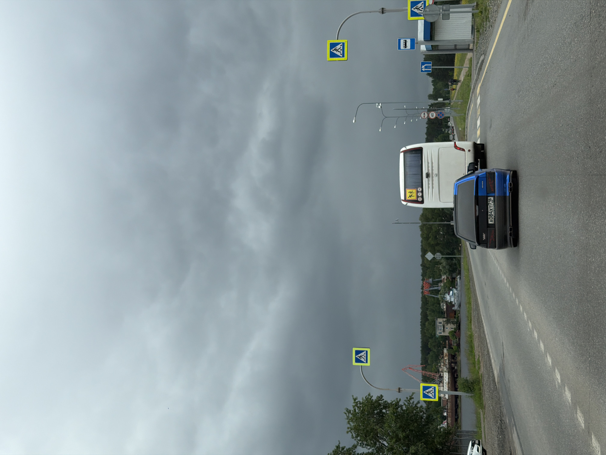 The image depicts an overcast, grey day on an asphalt road in Chaikovsky, Perm Krai, Russia. The sky is dominated by thick, dark grey clouds that cover the entire visible expanse. In the foreground, the rear of a black sedan, identified by the partial blue paint on its rear bumper and side, is visible, facing away from the viewer. The car bears the Russian license plate A084XT 716, which corresponds to the Perm Krai region. It features twin exhaust pipes. Ahead of this car is a white passenger bus, also facing away, with a yellow square sign depicting two black figures (indicating children) on its rear. Several other vehicles are visible further down the road in the distance, heading in the same direction. A small portion of a white car is parked off the road on the far left. The road features a dashed white line marking the center of the visible lanes and a solid yellow line delineating the right shoulder. A pedestrian crossing (zebra stripes) is visible further down the road. Multiple streetlights line both sides of the road. Various road signs are present: three yellow square signs with black pedestrian symbols (pedestrian crossing warnings) are visible – one on the left and two on the right. A blue square sign with a white bus symbol (bus stop) is located on the right, next to a light-colored bus stop shelter. Behind the bus, a circular red-rimmed sign indicating a 40 km/h speed limit and another circular prohibition sign are visible. A blue rectangular sign with two upward-pointing white arrows (indicating lanes) is also on the right. To the left of the road, beyond a strip of green foliage, lies a calm body of water, likely a river or reservoir. Along the far bank of this water body, industrial structures are visible. These include several large, orange-red gantry cranes and what appear to be multiple ships or boats, some seemingly derelict or under repair, docked or moored. A light-colored, multi-story building with numerous windows stands near the waterfront, amidst the industrial equipment. Dense green trees and other vegetation border both sides of the road and the distant bank of the waterway. On the right side of the road, past the bus stop shelter, two indistinct human figures are visible walking away from the viewer on a sidewalk or path. The roadside verges are covered with grass and some yellow