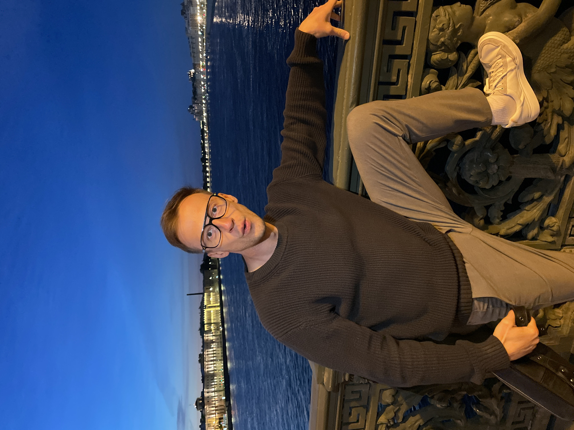 A man wearing glasses, a dark sweater, and light-colored pants is posing on a bridge at night. He is leaning on the bridge railing, with his right hand resting on it. His left leg is bent and his foot is resting on a decorative element of the bridge. He is holding a black case in his left hand. In the background, the Neva River is visible, with illuminated buildings and lights reflecting on the water. The sky is dark blue, indicating it is twilight or night. The architectural style of the buildings and the presence of the Neva River strongly suggest the location is Saint Petersburg, Russia. The bridge appears to be a historic one, given the ornate railings.