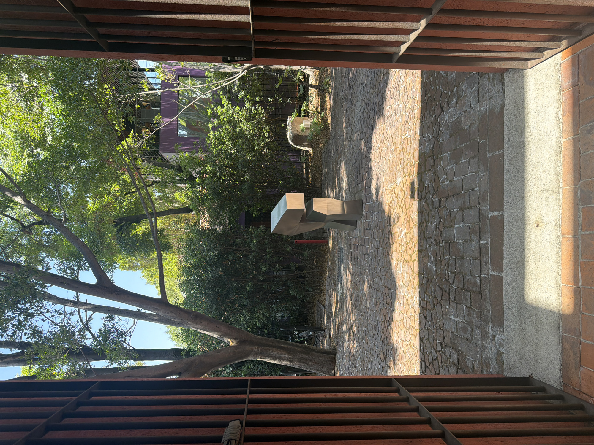 The image presents a view through a corridor-like structure with a reddish-brown metal framework. The view opens onto an outdoor space filled with greenery, featuring trees and a building with purple walls in the background. A modern, geometric sculpture made of light-colored material is placed in the center of the outdoor space. The ground is paved with stones or bricks. A bicycle is leaning against a tree. The light suggests it is a sunny day. The location is Puebla, Mexico.