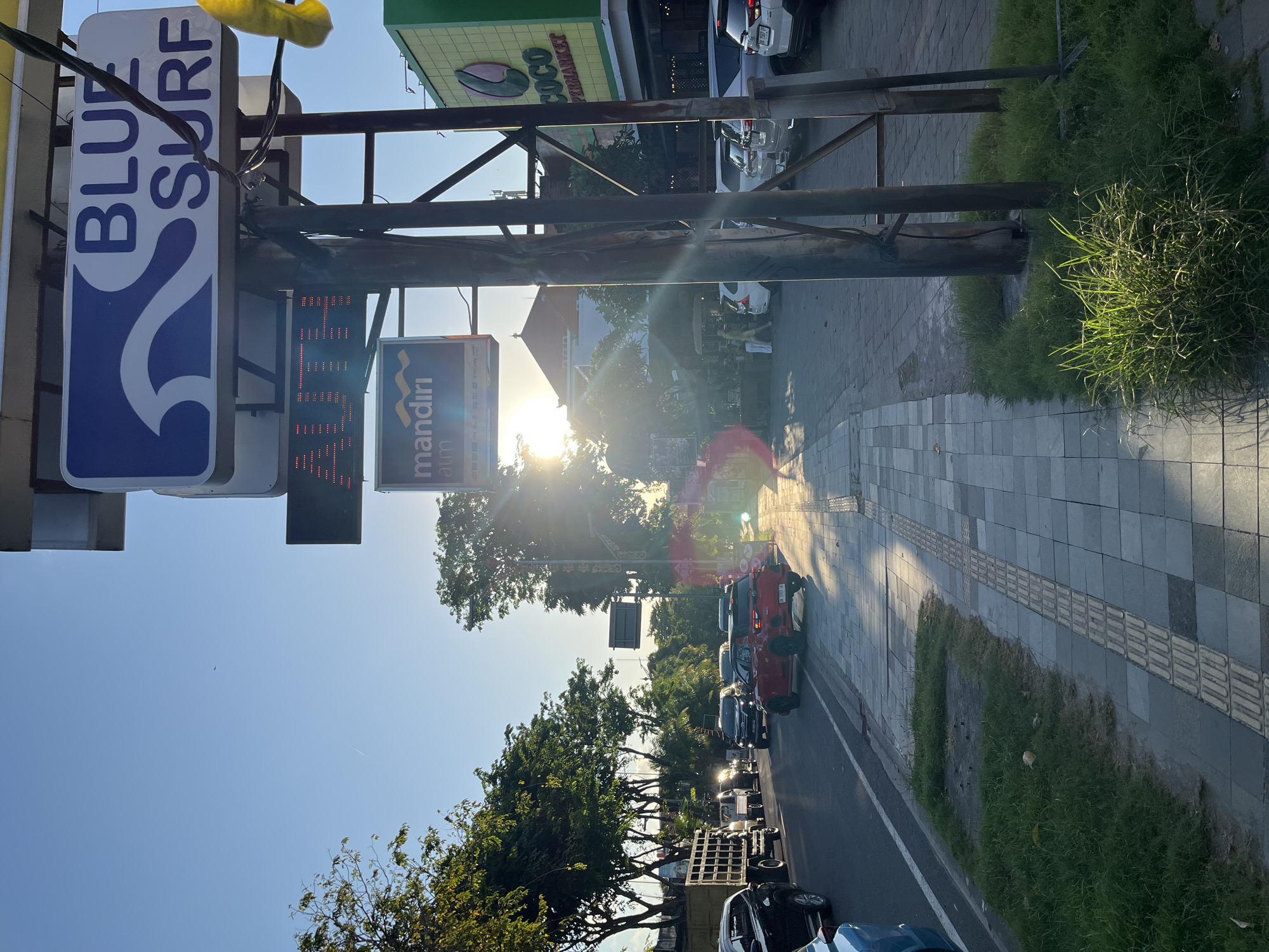 The image captures a street scene in Unknown, Indonesia. The sun, slightly obscured by trees, shines brightly down the road, casting a glare. Cars are parked along the roadside. A sidewalk runs parallel to the road, lined with a patch of grass. Visible signage includes "BLUE SURF," an ATM sign, and "COCO Supermarket."