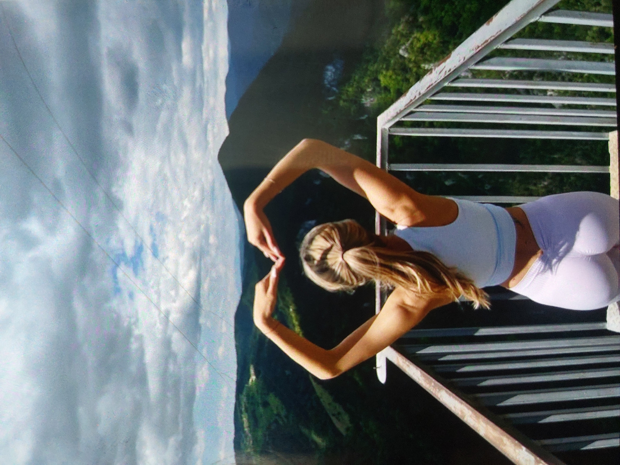 A young woman stands on a white metal balcony overlooking a mountainous landscape. She is wearing a light blue tank top and light purple leggings. The woman is holding her hands up to form a heart shape. Behind her is a panorama of green hills and a cloudy sky. It is unclear if this location is specifically within Tsarevo, Bulgaria, based on the image alone.