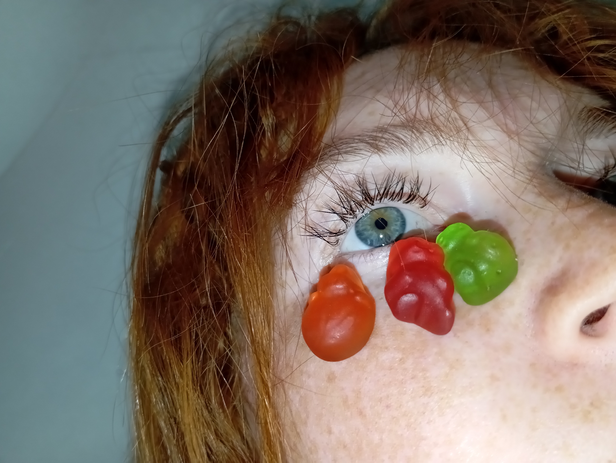 A close-up photograph depicts a portion of a person's face, specifically the left eye and adjacent areas. The person has fair skin with freckles and light-colored eyelashes. The eye is blue. Three gummy bears of different colors (orange, red, and green) are placed on the person's face below the eye. The person has reddish-brown hair. The background is a neutral color, likely a wall. The scene is not identifiable as being in Yurgamysh, Russia.