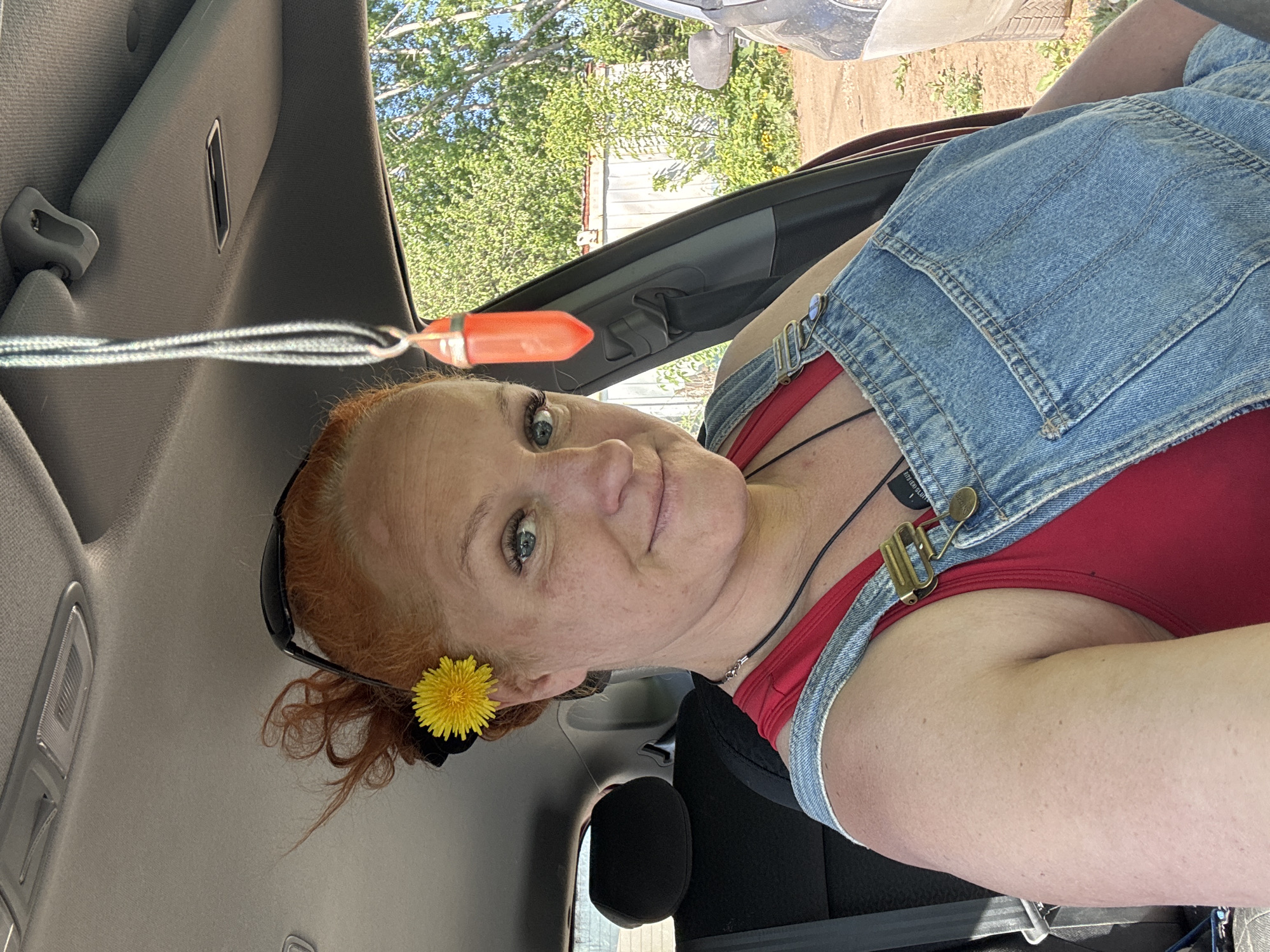 The image shows a woman with red hair wearing a red shirt and denim overalls, sitting in a car. A yellow flower is tucked behind her ear. The car's interior is visible, including the headrest and seatbelt. Outside the car window, there are trees and a building visible. The presence of the city of Chaikovsky, Russia, cannot be confirmed in the image.