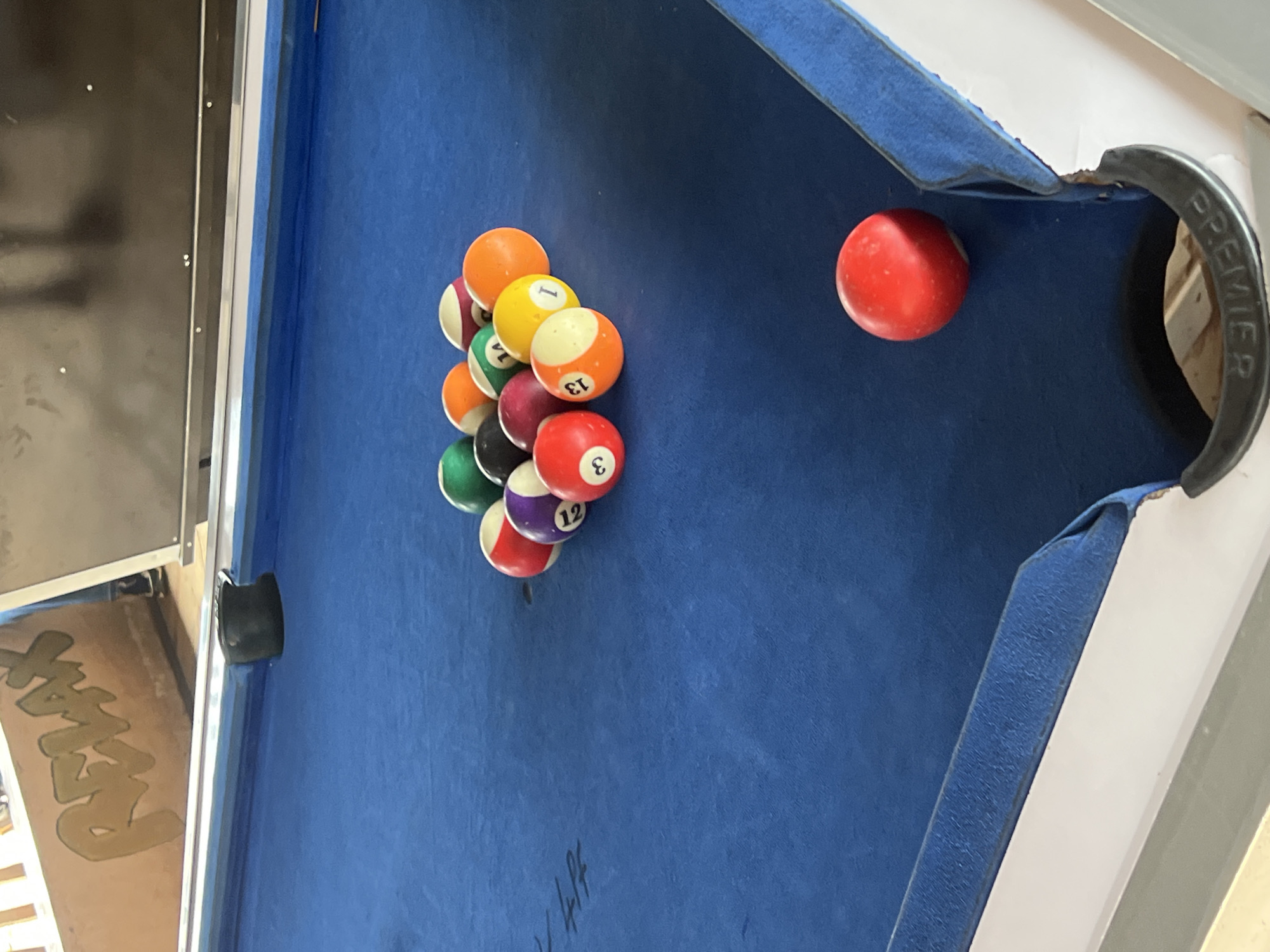 The image shows a close-up view of a blue billiard table with a rack of pool balls. The balls are arranged in a triangular formation, with a red ball in the foreground to the right of the frame. Some of the balls have numbers and colors visible, including a red ball with the number 3, an orange and white ball with the number 13, a yellow ball with the number 1, a purple ball with the number 12, and a green ball with the number 14. The background shows a wall with the word "RELAX" painted in large, stylized letters. The overall scene appears to be in an indoor setting, likely a bar or recreational facility. There is no indication of the location being in Durimi, Nigeria.
