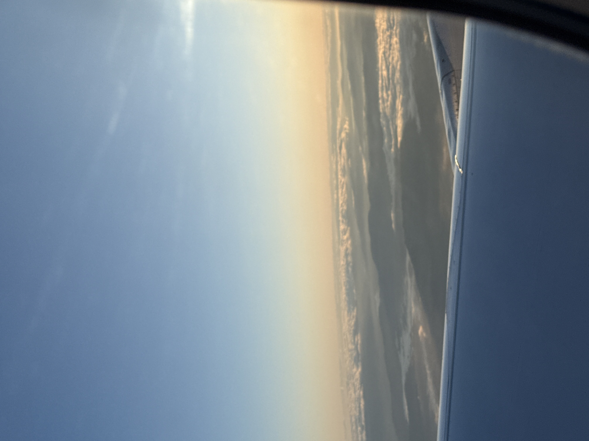 The image shows a view from an airplane window. The sky is visible, transitioning from blue to a hazy yellow-orange color. Below the horizon line, there appear to be either clouds or land formations partially obscured by haze. A portion of the airplane wing is visible in the lower right of the frame. The location is unlikely to be identified as Moreira, Portugal from this image, because of a lack of specific identifiable landmarks.