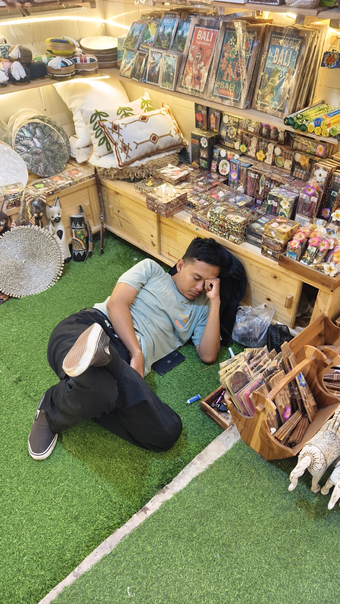 The image shows a young man sleeping on artificial green turf inside what appears to be a gift shop. The shop is located in Pecatu, Indonesia, as indicated by the "Bali" and "Indonesia" tourist memorabilia on the shelves behind him. The man is lying on his side with his head resting on a black bag. He is wearing a light blue t-shirt, black pants, and black sneakers with one foot raised slightly. His cell phone and a blue pen are on the turf near him. The shelves behind the man display a variety of souvenirs, including postcards, incense, carved wooden items, and small trinkets arranged in boxes. There are also decorative pillows, woven baskets, and small wooden sculptures visible. The shop is well-lit, and the overall scene suggests a quiet moment amidst the merchandise.