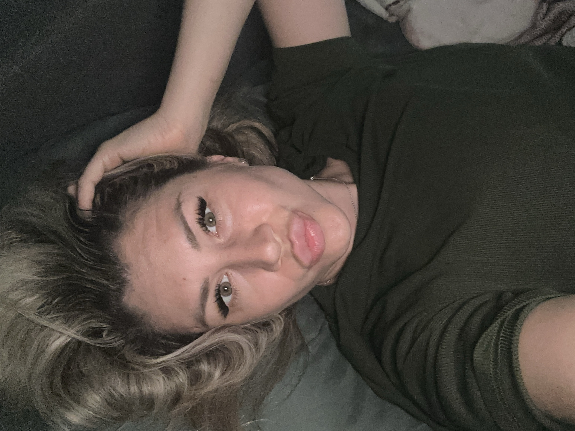 A woman with blonde hair and green eyes is lying on her back, looking up at the camera. She is wearing a dark green shirt and has her hand on her head. Her lips are pursed. The background appears to be a dark, textured surface, possibly a couch or bed. The image was taken in Novoaltaysk, Russia.