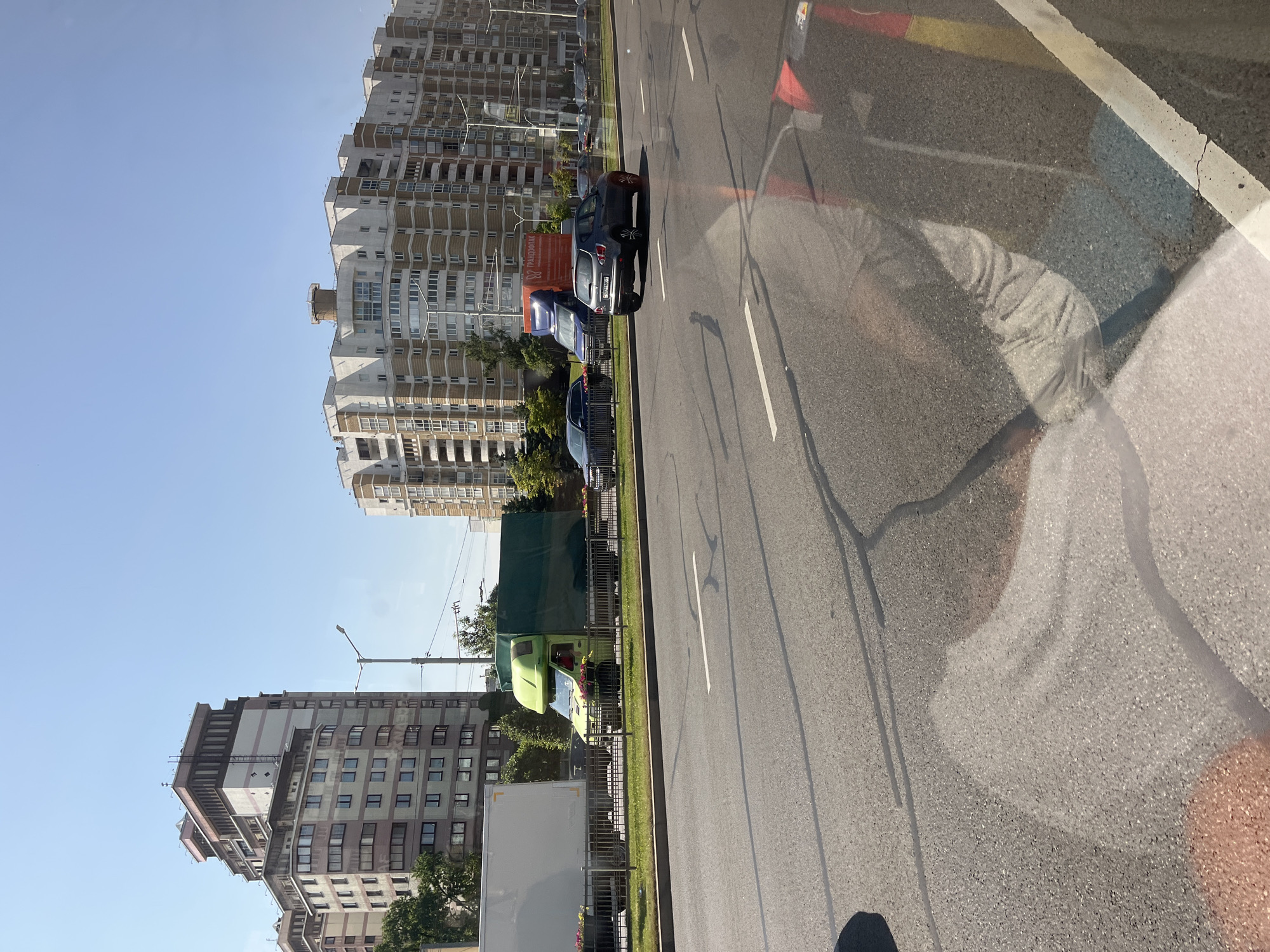 The image depicts a street scene in Minsk, Belarus. In the background, modern apartment buildings with cream and brown facades dominate the skyline. The buildings feature numerous balconies and windows. A clear blue sky is visible above. In the middle ground, a multi-lane road stretches across the frame. Several vehicles are present on the road and parked on the side. These include a white delivery truck, a bright green truck with a covered cargo area, a blue truck with an orange advertisement panel, a dark gray hatchback car, and a dark blue sedan. A metal fence separates the road from a grassy median where trees and streetlights are visible. The foreground of the image shows the asphalt road surface with visible lane markings and reflections. The reflections on the road surface are from the viewer's perspective, suggesting the image was taken from inside a vehicle.