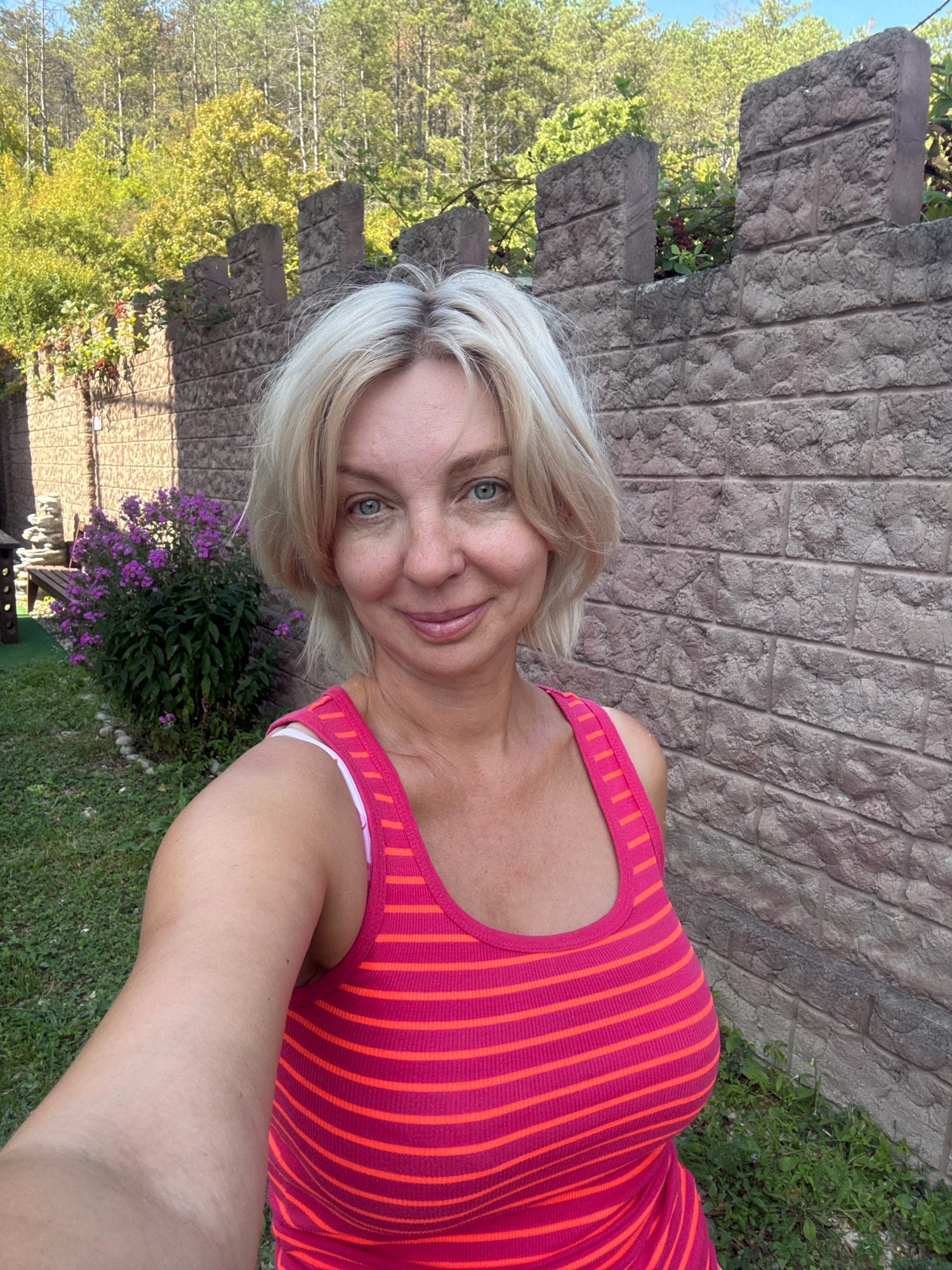 A middle-aged Caucasian woman with blonde hair and blue eyes is taking a selfie outdoors. She is wearing a pink and orange striped tank top. She is standing in front of a stone wall and a grassy area with some purple flowers. In the background, there are trees. The setting appears to be a garden or yard, possibly in Abrau-Dyurso, Russia.