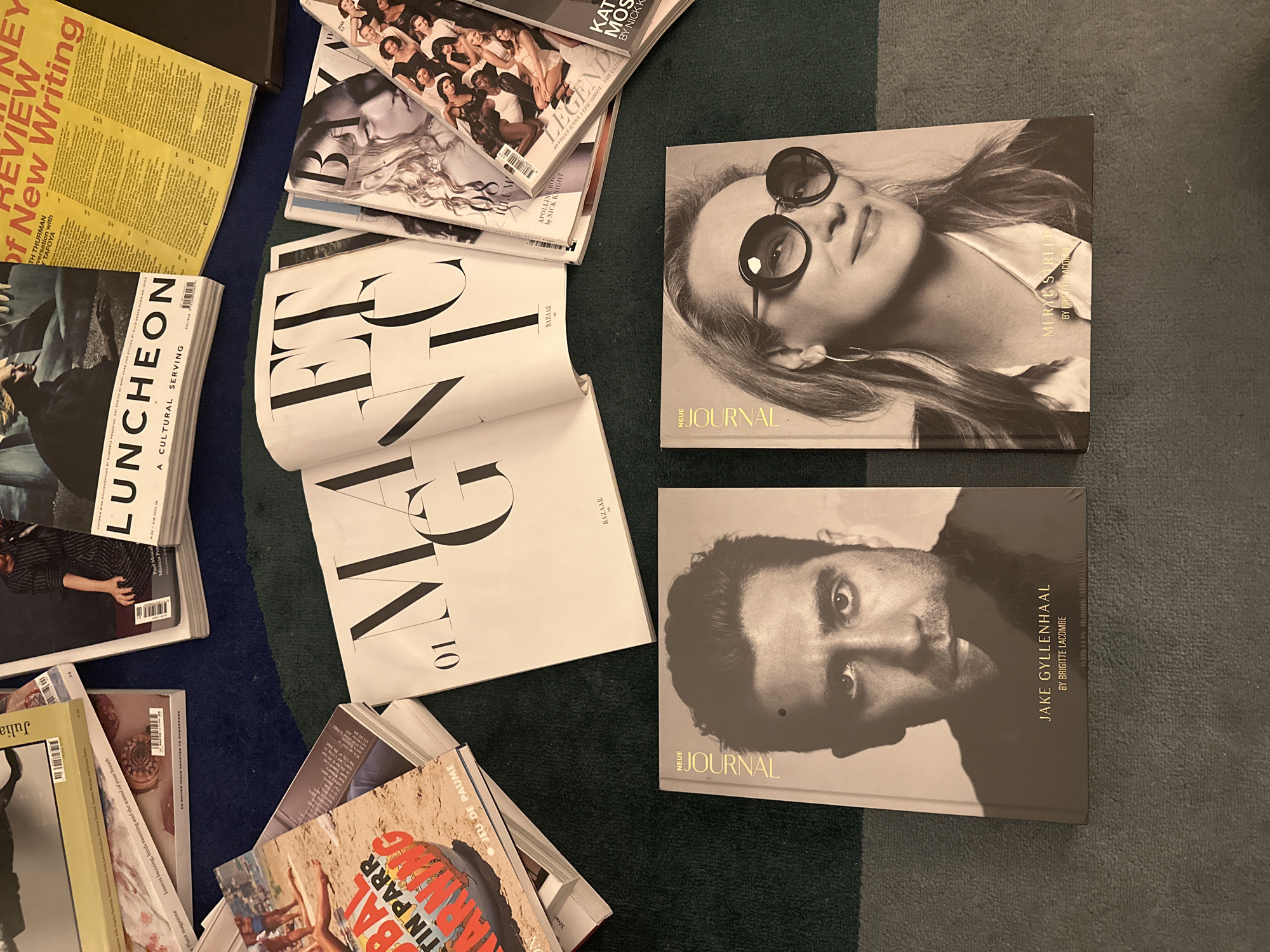 The image shows a collection of magazines and books scattered on a green and blue striped carpet. The magazines include "Luncheon: A Cultural Serving," "Bazaar," "The Journal," "Plant Magazine," and others. One "The Journal" book features a black and white portrait of Meryl Streep, while another shows Jake Gyllenhaal. One partially visible magazine has the text "BAL" "ARNING" "TIN PARR" JEU DE PAUME. The environment is otherwise unidentifiable. There are no people visible in the image.