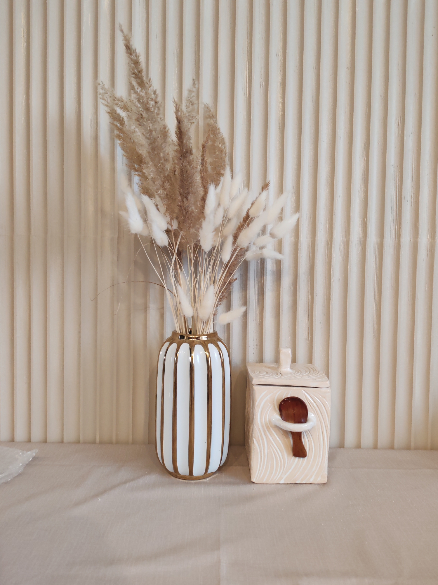 The image displays a serene indoor still life arrangement, likely in Ust-Ilimsk, Russia, featuring two decorative objects against a textured wall. In the left-center, a tall, elegant white ceramic vase with vertical gold stripes holds a bouquet of dried grasses. The bouquet comprises both fluffy, light brown pampas grass and softer, off-white bunny tail grass, creating a natural and airy aesthetic. To the right of the vase, a squat, light peach-colored ceramic container is positioned. This container has a square base and a distinctive, house-like lid with a small handle on top. A small, dark brown wooden spoon hangs from a decorative, sculpted loop on the container's front. The surface of the container features a subtle, wavy relief pattern. Both items rest on a light beige or off-white fabric surface, possibly a tablecloth, which extends across the foreground. The background is a prominent wall covered in vertical, ribbed panels, painted in a matching light beige tone, providing a consistent and understated backdrop. The lighting is soft and even, suggesting an indoor daytime setting. There are no people visible, and no text is discernible in the image. The overall impression is one of modern, minimalist decor.