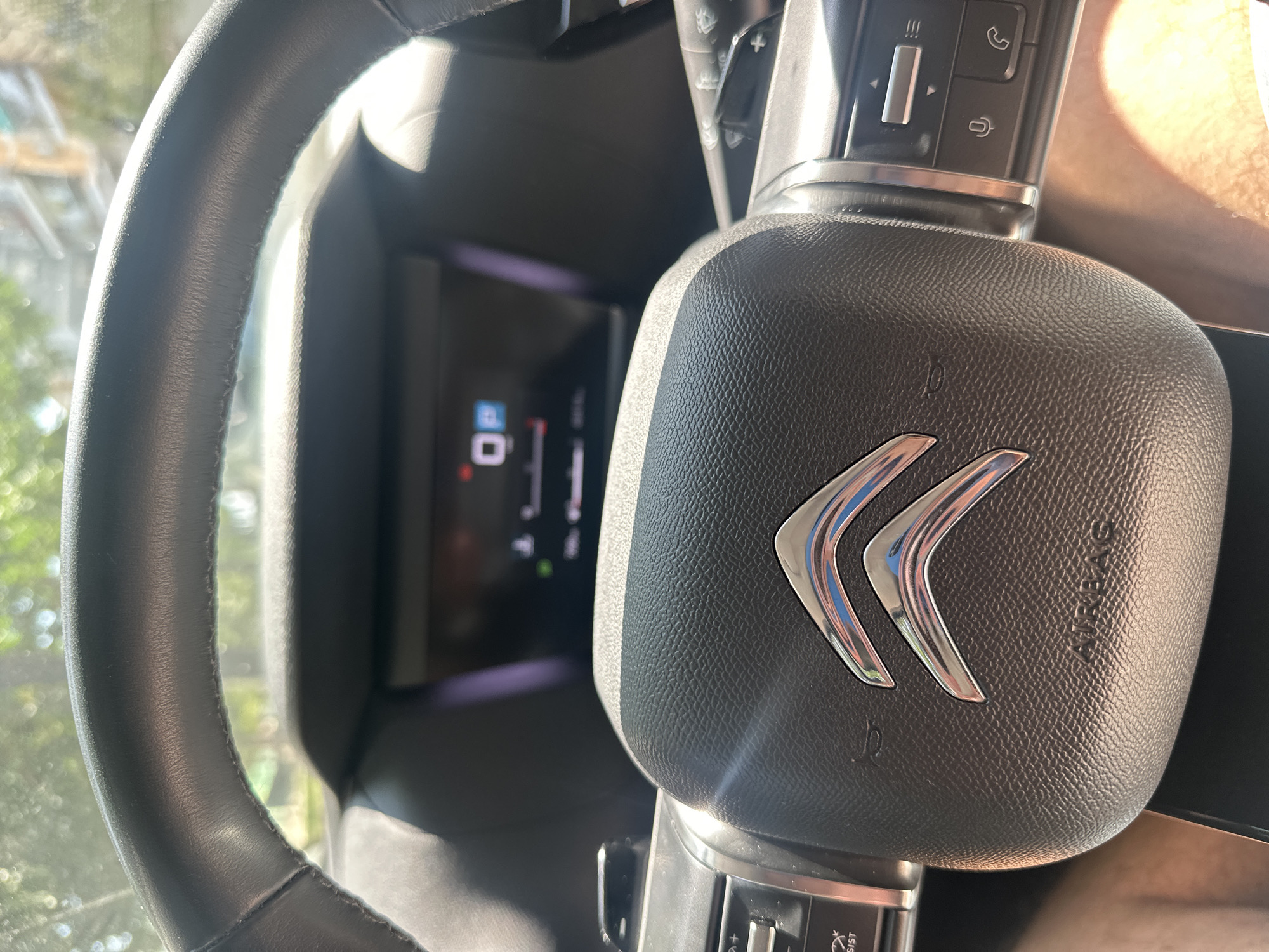 The media file is a close-up, eye-level shot of the interior of a car, specifically focusing on the steering wheel and dashboard. The steering wheel is black and appears to be made of leather, with stitching visible along its rim. In the center of the steering wheel is a silver emblem consisting of two chevrons, the logo of the car manufacturer, Citroën. The word "AIRBAG" is embossed on the steering wheel's central pad.
To the left of the steering wheel, a portion of the car's digital dashboard is visible. It displays a digital gauge with a reading of "0" and a lock symbol, along with other indicators that are partially obscured. On the right side of the steering wheel column, there are several control buttons, including what appear to be volume controls and buttons with icons for a phone and a cruise control or speed limiter system.
The scene is brightly lit, suggesting it is daytime. Sunlight is casting reflections on the steering wheel and dashboard, and a warm, hazy light is visible in the upper right portion of the frame, possibly from sunlight entering through the windshield or window. The background is blurred, showing hints of green foliage and structures outside the car, indicating it is parked outdoors. The overall impression is a snapshot of a car's interior on a sunny day in what is identified as Benimàmet-beniferri, Spain. No people are visible in the image.