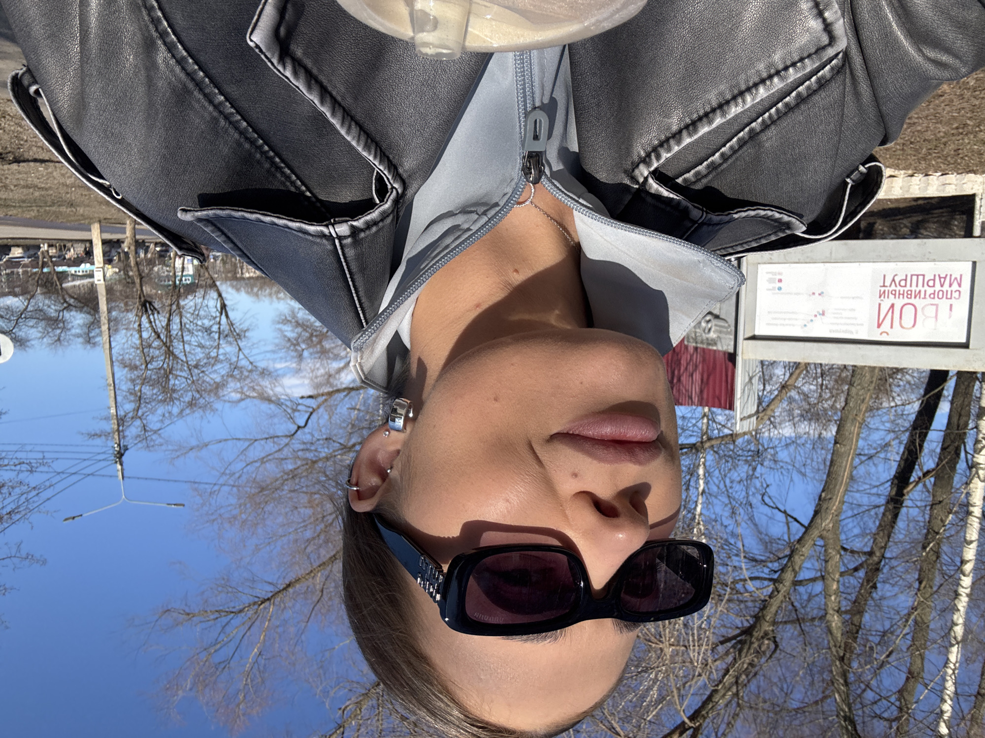 A person with fair skin, wearing black sunglasses, multiple earrings in the left ear, a gray jacket, and a gray hooded shirt, is positioned outdoors on a sunny day. The background includes bare trees and a clear blue sky. A sign with Russian text, including the word "СПОРТИВНЫЙ" (sportivnyy, meaning "sporting" or "athletic") and "МАРШРУТ" (marshrut, meaning "route"), is visible. The presence of the Russian text suggests a location in Russia. Chernushka is identified as the city location. The orientation of the image is tilted, and the person is looking down towards the camera. Part of a clear plastic container is visible in the upper part of the image.