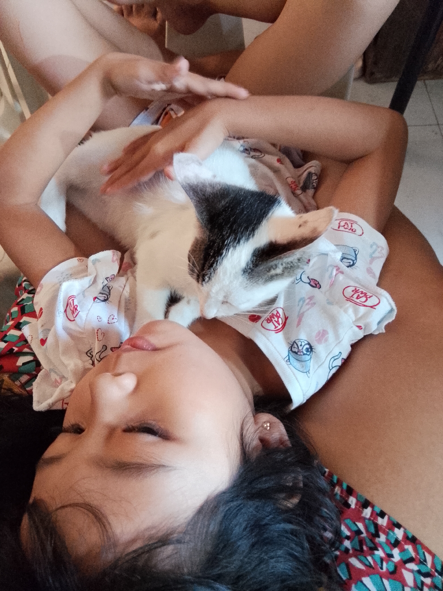 This media file captures a peaceful indoor scene, likely a home in Davao City, Philippines, featuring a young child and a cat sharing a moment of quiet companionship. The subject is a child, appearing to be a girl, lying down, seen from the chest up, with her eyes closed or partially closed, suggesting sleep or deep relaxation. She wears a white short-sleeved top adorned with playful cartoon cat designs, including a cat's face, the word "CAT" within a speech bubble, and the initials "WR." A small, gold-colored stud earring is visible in her left ear.
A black and white cat is resting comfortably on the child's chest or stomach area, its head gently placed near the child's face. The cat also appears to be wearing a similar white garment, possibly a custom onesie, patterned with cartoon cat illustrations and the word "CAT." The child's hands are softly resting on or near the cat, underscoring their affectionate bond.
In the background, the legs or thighs of another individual, presumably an adult, are partially visible, hinting at their presence nearby. A dark chair leg is also seen in the upper right portion of the image. The flooring beneath them appears to be tiled, a common feature in homes in the Philippines. The soft, natural lighting suggests it is daytime. The overall atmosphere is one of warmth, tranquility, and a loving connection between the child and her pet.
