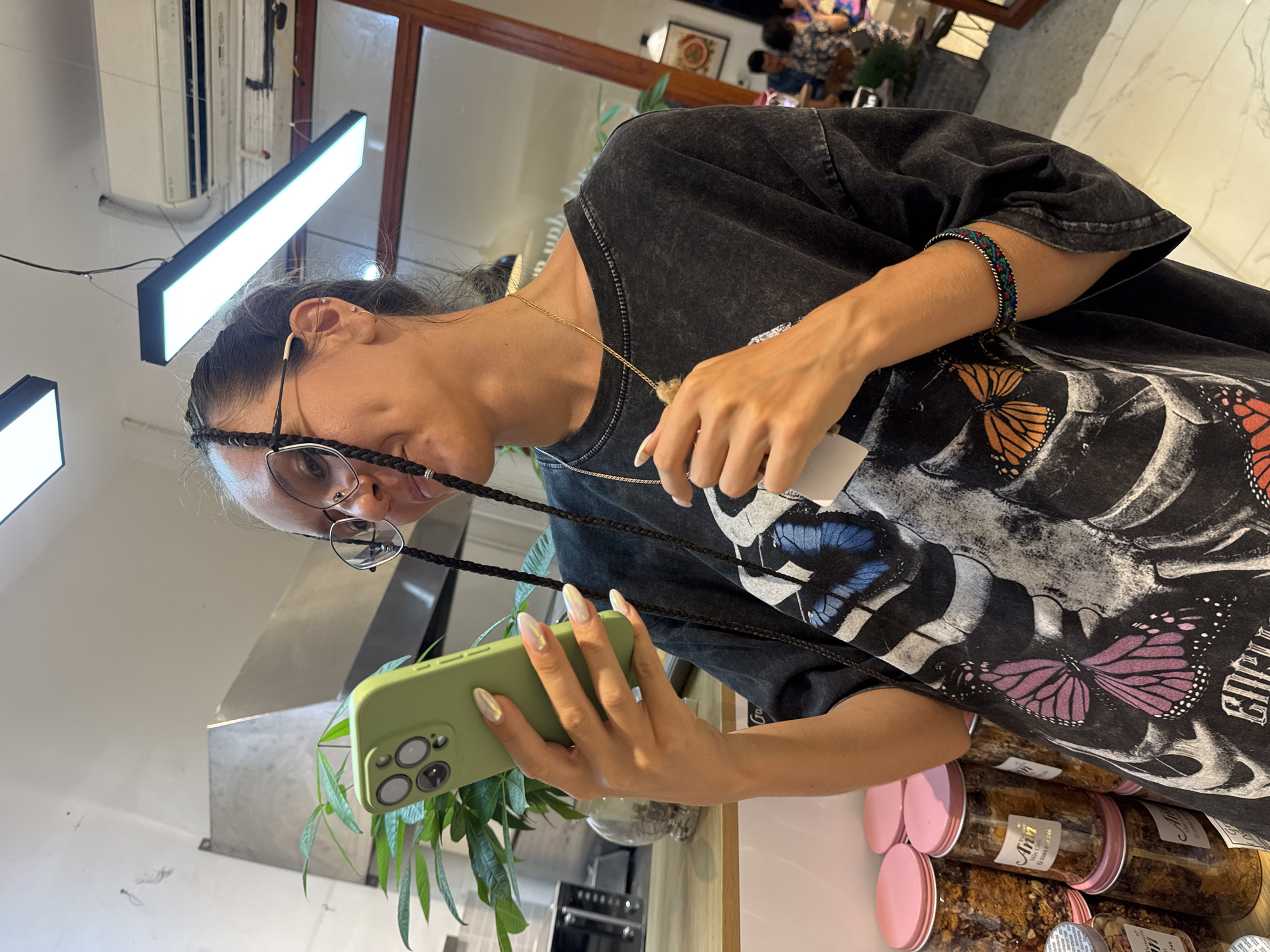 A young woman with dark hair in braids and round glasses is looking at her light green smartphone in Mộc Châu, Vietnam. She is wearing a dark, oversized t-shirt with a skeletal design and butterflies. She has a gold necklace and a colorful bracelet on her wrist. In the background, there are jars of food on shelves, a plant, and a kitchen exhaust hood. The scene appears to be a cafe or restaurant.