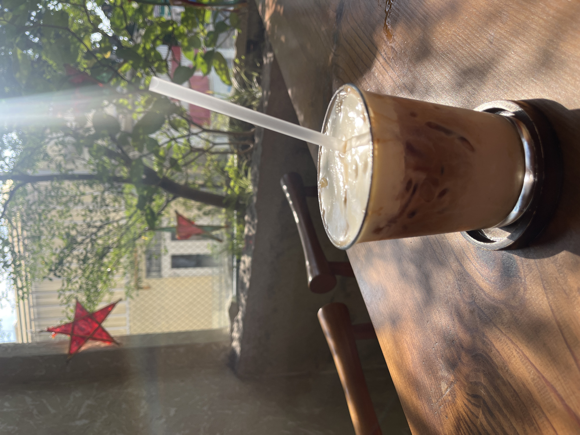 This image depicts an iced coffee drink placed on a wooden table, with a straw inserted into it. The drink is in a clear glass with some condensation visible. The table has a rich, dark wood grain. In the background, sunlight streams through a window, illuminating greenery and what appear to be two red star decorations hanging by the window. The overall setting suggests an indoor cafe or restaurant with a view of an outdoor area. The lighting and the presence of greenery hint at a bright, possibly tropical, environment. The context of Ho Chi Minh City, Vietnam, suggests this could be a cafe in a bustling urban area, perhaps with a relaxed, outdoor-feeling atmosphere, possibly during daylight hours. The image focuses on the drink, creating a sense of a quiet moment enjoyed in a cafe.
