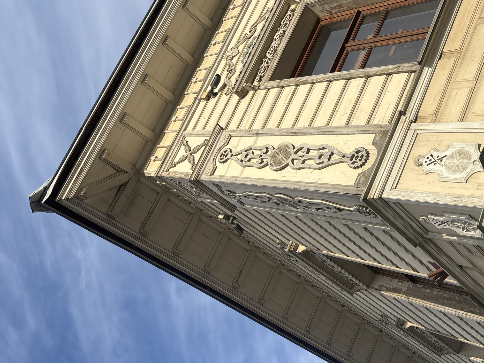 The image shows a low-angle view of a decorative, wooden building exterior against a blue sky. The building has light yellow/cream-colored siding with ornate, carved wooden trim in a lighter, grayish tone. A window with a brown frame and multiple panes is visible. The angle of the photo suggests the building is viewed from below. The building is the Meshkov House, located in Perm, Russia.