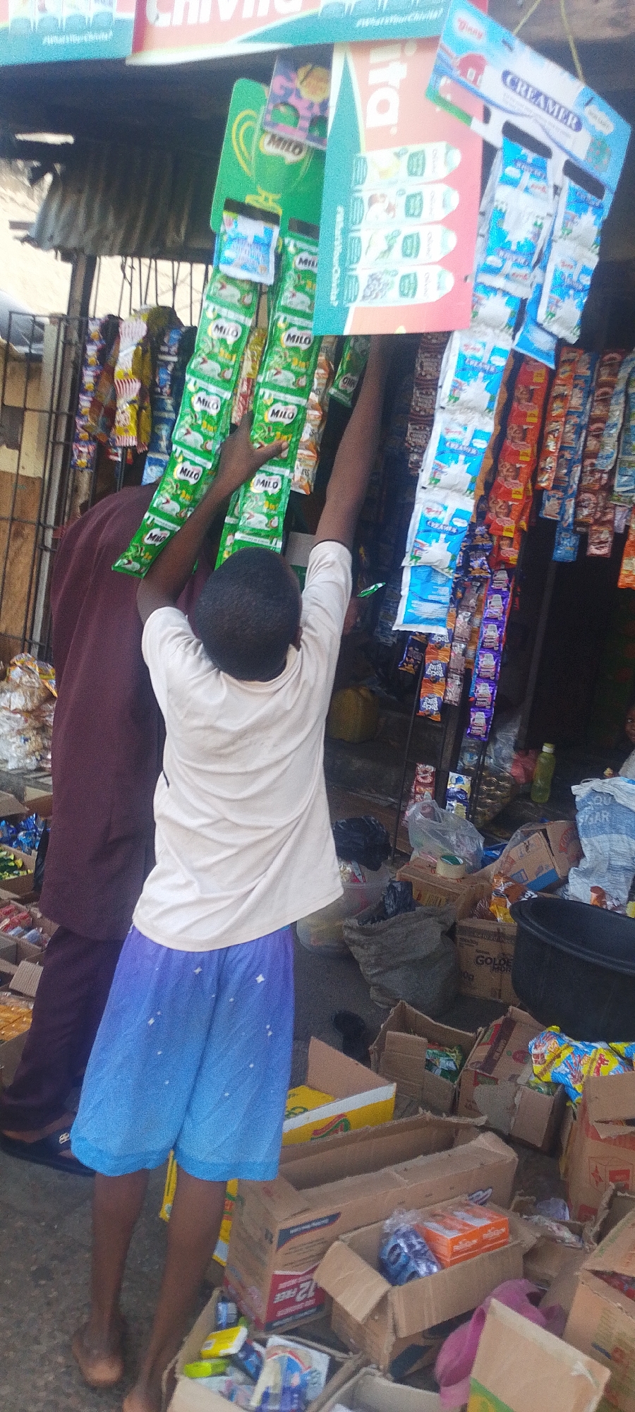 A view of a vendor's makeshift kiosk in Lokoja, Nigeria, constructed with corrugated metal. The kiosk is stocked with small packets of Milo, creamer, and other products hanging from the front. A young boy with blue shorts is reaching for the Milo packets, and another person in a maroon garment stands beside him. The ground around the kiosk is cluttered with boxes of merchandise.