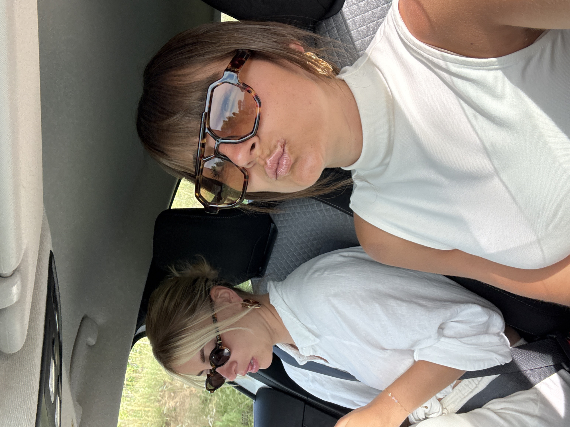 Two women are sitting inside a car. The woman in the foreground is wearing a white tank top and large brown-framed sunglasses. She has short brown hair and plump lips. She is facing the camera with a slight pout. The woman in the background is wearing a white button-down shirt and small, round sunglasses. Her blonde hair is pulled back in a bun. She is looking down and appears to be wearing a seatbelt. The interior of the car is visible, including the dashboard, seats, and headrests. The background outside the car appears to be a natural environment with trees and greenery. The location is possibly Gassin, France, due to the landscape, though that isn't determinable with certainty from the image.