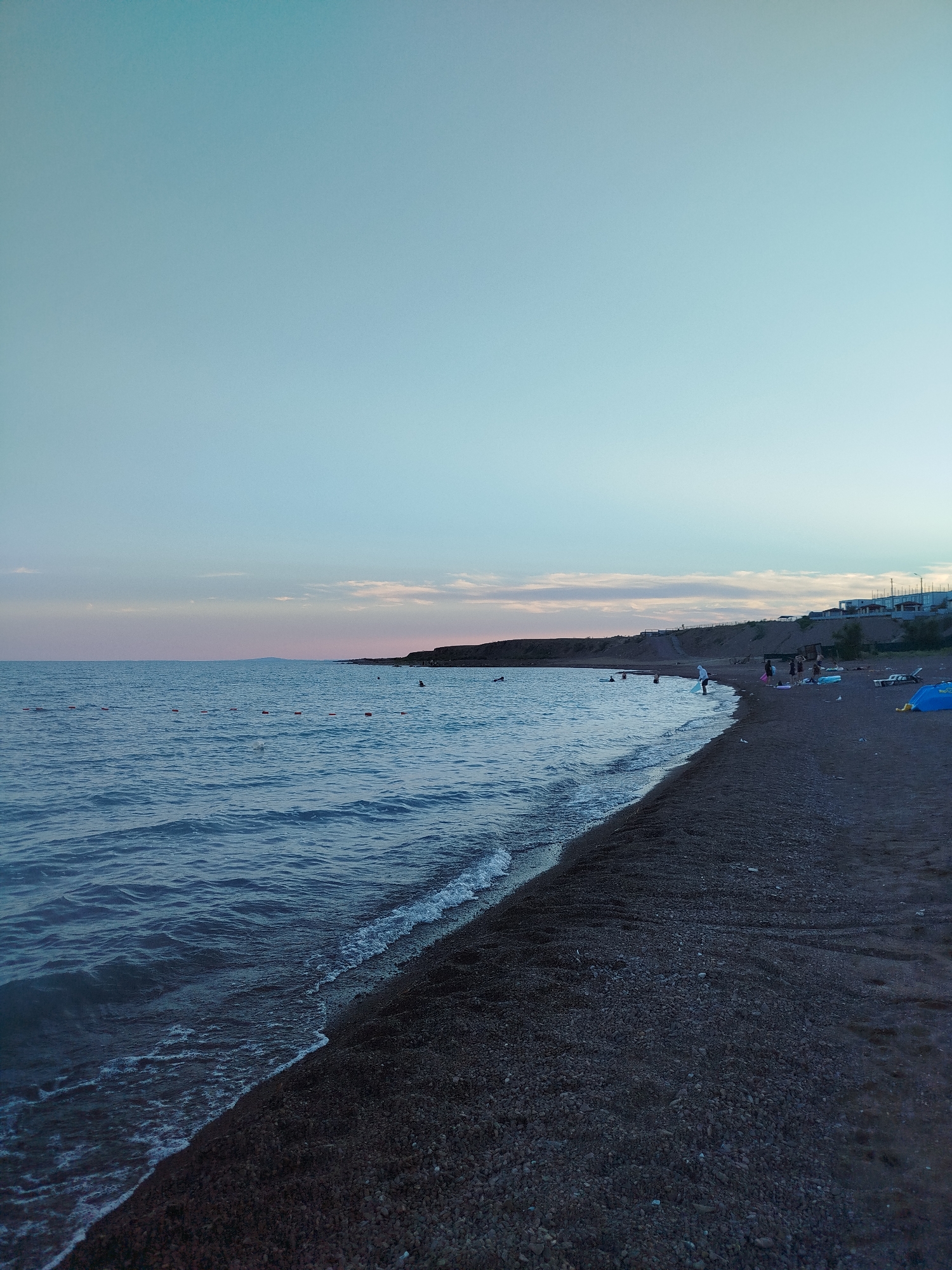 The image depicts a serene beach scene at dusk. The sky is a gradient of soft blues and pale pinks, with a few wispy clouds near the horizon. The sea is a deep blue, with gentle waves lapping at the shore. The beach is made up of dark pebbles and sand, and the water's edge is marked by a thin line of white foam. In the distance, a line of buildings and lights suggest a settlement along the coast. Scattered along the shoreline are a few small groups of people, some appearing to be wading in the water, while others are standing or sitting on the beach. One person is standing in the water near the shore, appearing to interact with something. A bright blue tent is visible on the beach, along with a beach chair. The overall atmosphere is tranquil and peaceful, with the fading light casting a soft glow over the scene. The specific location within the city of Unknown, Unknown cannot be determined from the visual information alone.