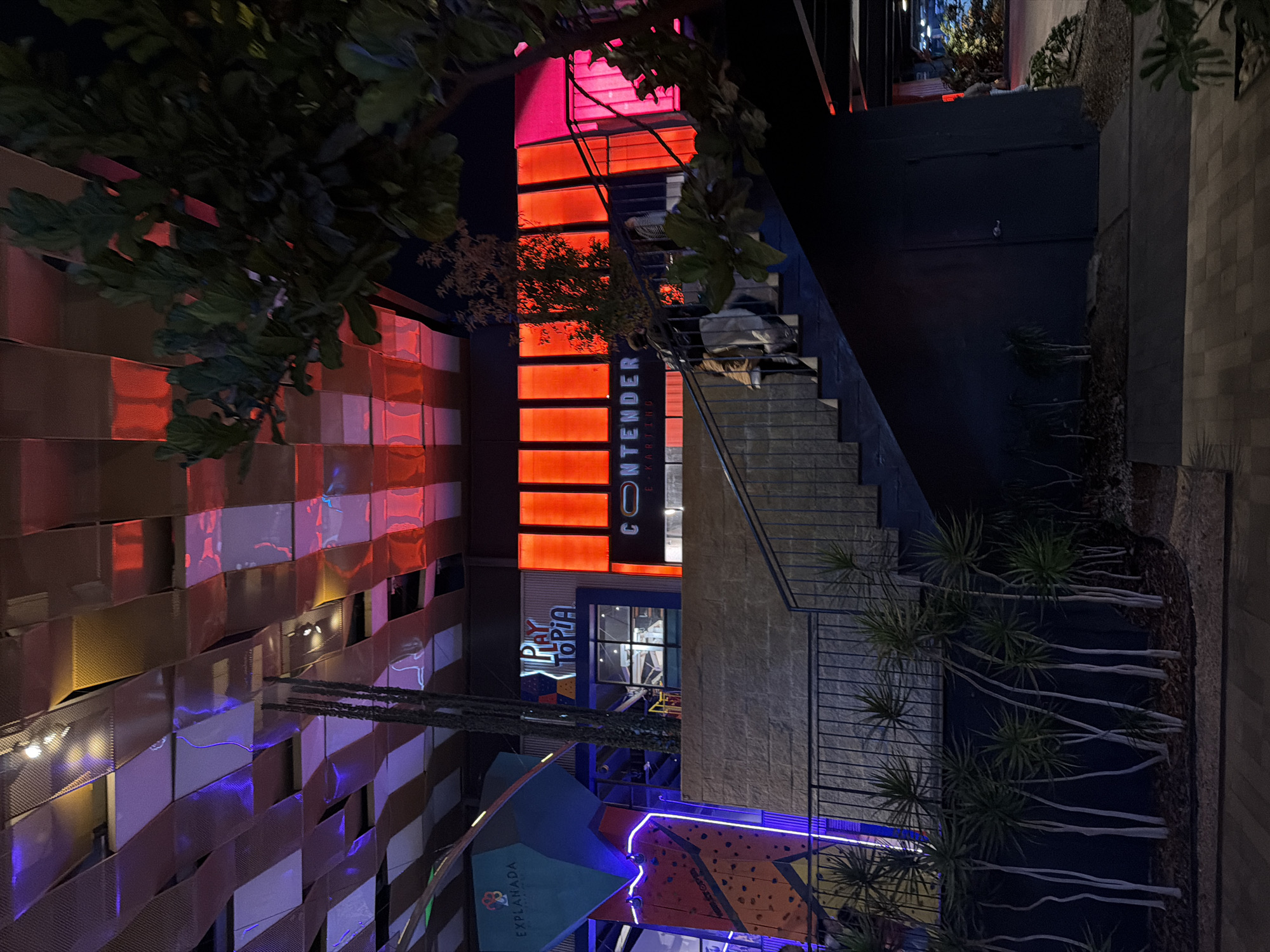 The image captures a nighttime scene in San Pedro Cholula, Mexico, featuring an exterior view of a complex containing several businesses and recreational areas. The building has a modern design with a mix of materials, including a lattice-like wall on the left illuminated with alternating red and purple lights. A prominent red sign with the word "CONTENDER" vertically displayed identifies a business. Stairs lead up to an entrance above. Below, a sign reading "PLAYTOPIA" is visible, and a brightly lit climbing wall with blue and orange sections is positioned at the bottom. The surrounding environment includes greenery along the stairs and a sidewalk leading into the frame on the right.