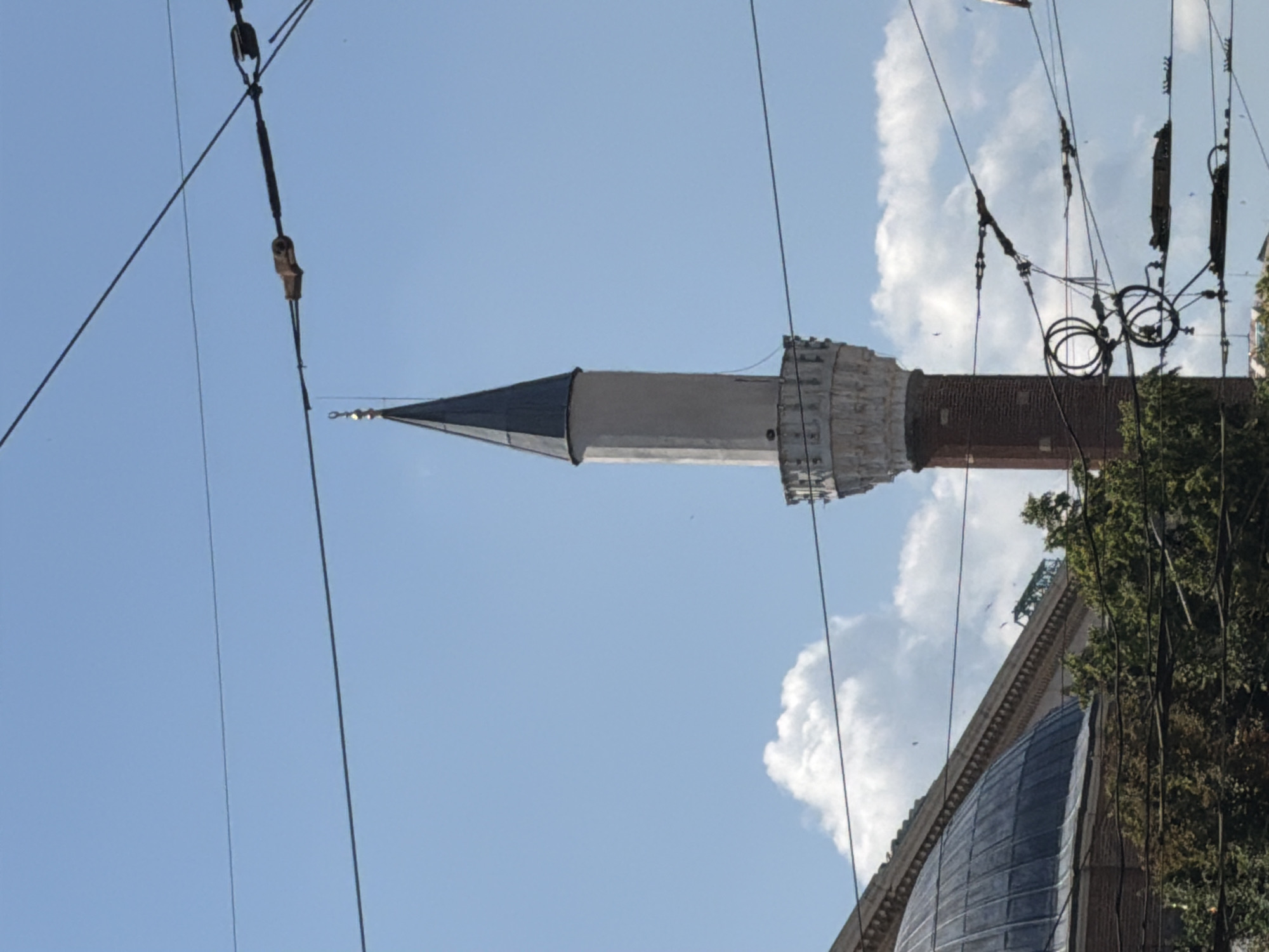 A minaret, part of a mosque, rises against a bright blue sky with scattered white clouds. The minaret features a red brick base, a white cylindrical shaft, and a dark conical top adorned with a gold finial. Below the minaret, the domed roof of a building and green trees are visible. Overhead, a network of electrical wires crisscrosses the frame, with several insulators and tensioners visible. The scene is likely located in Sofia, Bulgaria, given the architectural style and presence of overhead wires which are common in the city's public transport infrastructure.