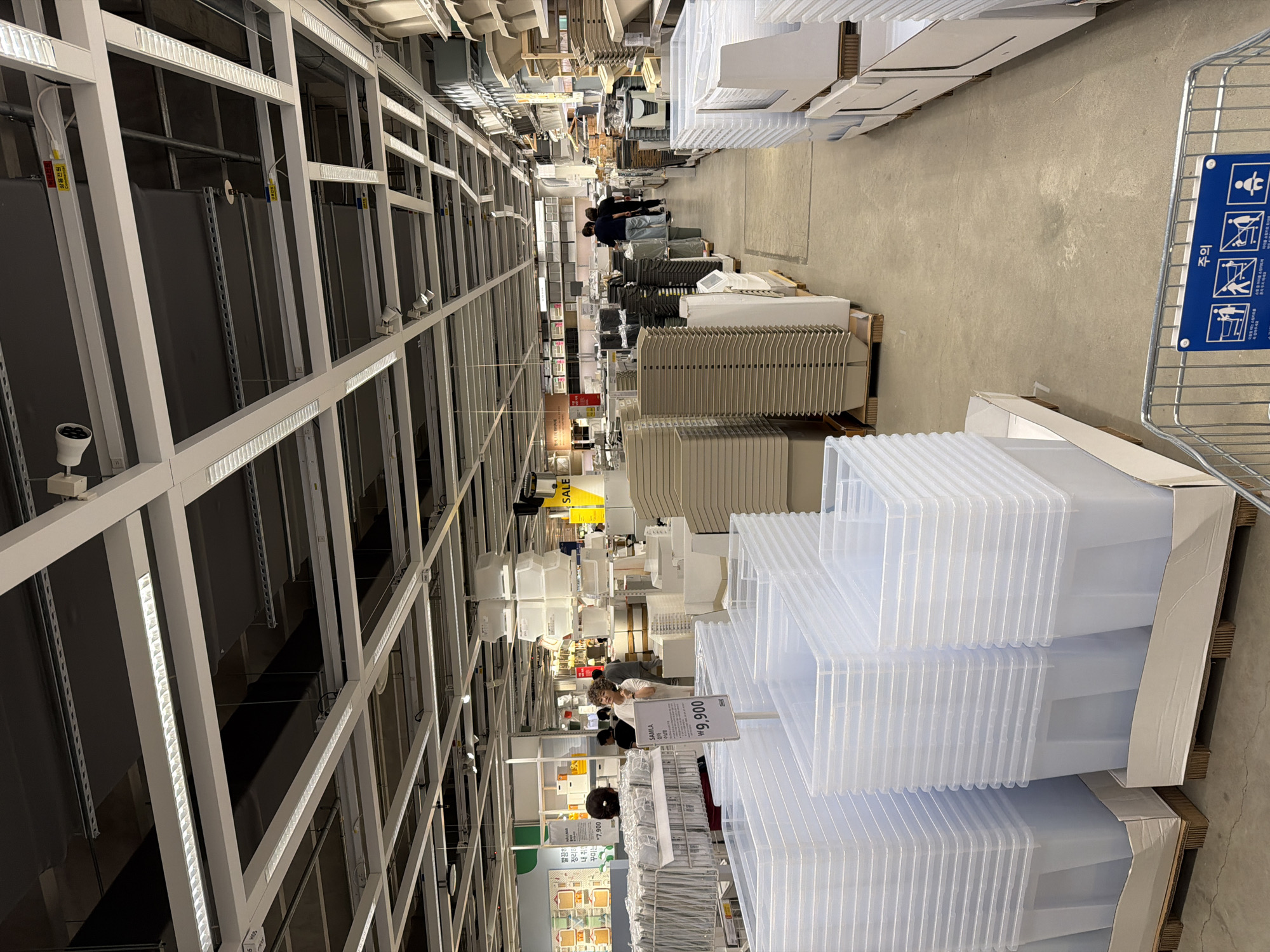 This is an indoor shot of a retail store, likely IKEA, located in Goyang-si, South Korea, based on the Korean signage visible. The scene depicts a wide aisle in a home goods store, with numerous storage containers, shelves, and display racks. Fluorescent lights are mounted on a grid-like ceiling structure. In the foreground, large, clear plastic storage bins are stacked on pallets. To the left, more rows of storage containers are visible, along with shelving units displaying various items. In the middle ground, a woman with curly hair is looking upwards, seemingly at a display or product. Further down the aisle, two individuals are walking away from the camera, and more shelves and storage solutions are apparent. A sign with Korean text is visible, and another sign in the foreground reads "₩9,900", indicating pricing. The floor is concrete. The overall atmosphere is that of a busy retail environment focused on home organization products.