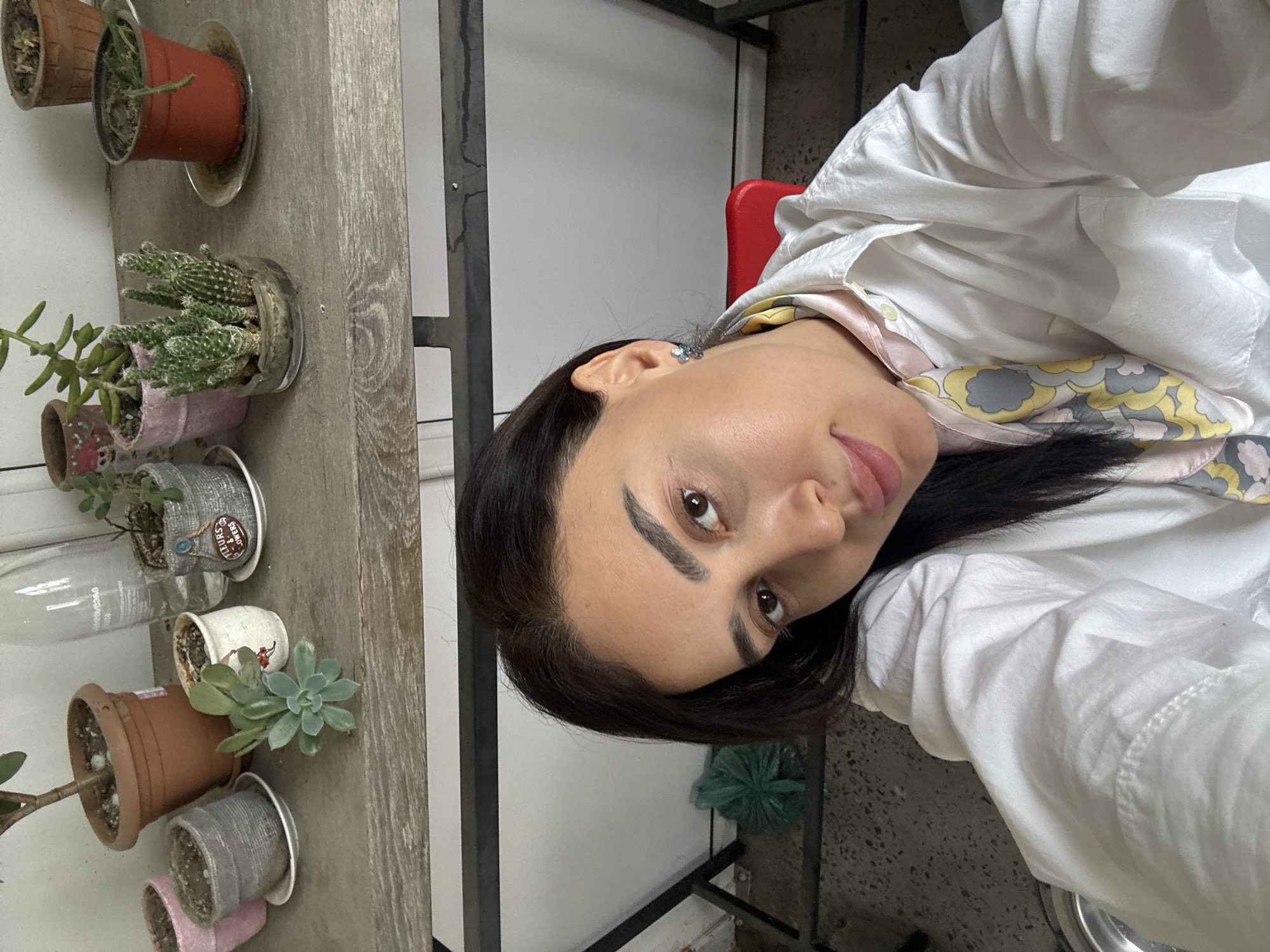 A woman with dark hair and brown eyes is facing the camera. She is wearing a white shirt over a patterned scarf. Behind her, a wooden structure holds various potted plants, including cacti and succulents. The indoor location suggests a residential or commercial space. The exact location within Tbilisi, Georgia is not discernible from the image.