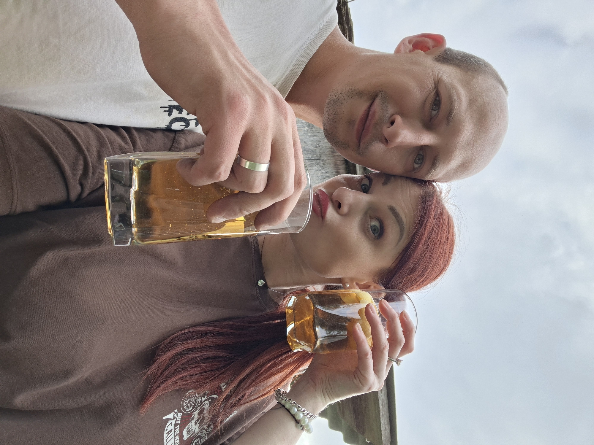 The image depicts two individuals, an adult male and an adult female, engaged in a close-up, low-angle selfie outdoors in Забалоцкі сельскі Савет, Belarus.
The male, positioned on the left, has a light complexion, a receding hairline/bald scalp, and light-colored eyes. He has short stubble on his face and is smiling towards the camera. He wears a plain white t-shirt and a silver-colored band ring on his left ring finger. He holds a clear, cylindrical glass containing an amber liquid.
The female, positioned on the right, has long red hair, a light complexion, and light-colored eyes. She is making a pouting or "duck face" expression with red lipstick. She wears a brown t-shirt with a partially visible graphic print that includes text. Her accessories include pearl stud earrings, a delicate silver-colored necklace with a small circular pendant, a pearl and chain bracelet on her left wrist, and a ring on her right ring finger. She holds a clear glass with an amber liquid and a visible lemon wedge.
Both individuals are holding their respective glasses towards the camera, suggesting a toast or a shared drink. The background is blurred but shows an overcast sky with light grey clouds and a weathered wooden structure, possibly a fence or building, indicating an outdoor, possibly rural or semi-rural, setting. The lighting is diffuse, consistent with an overcast day.