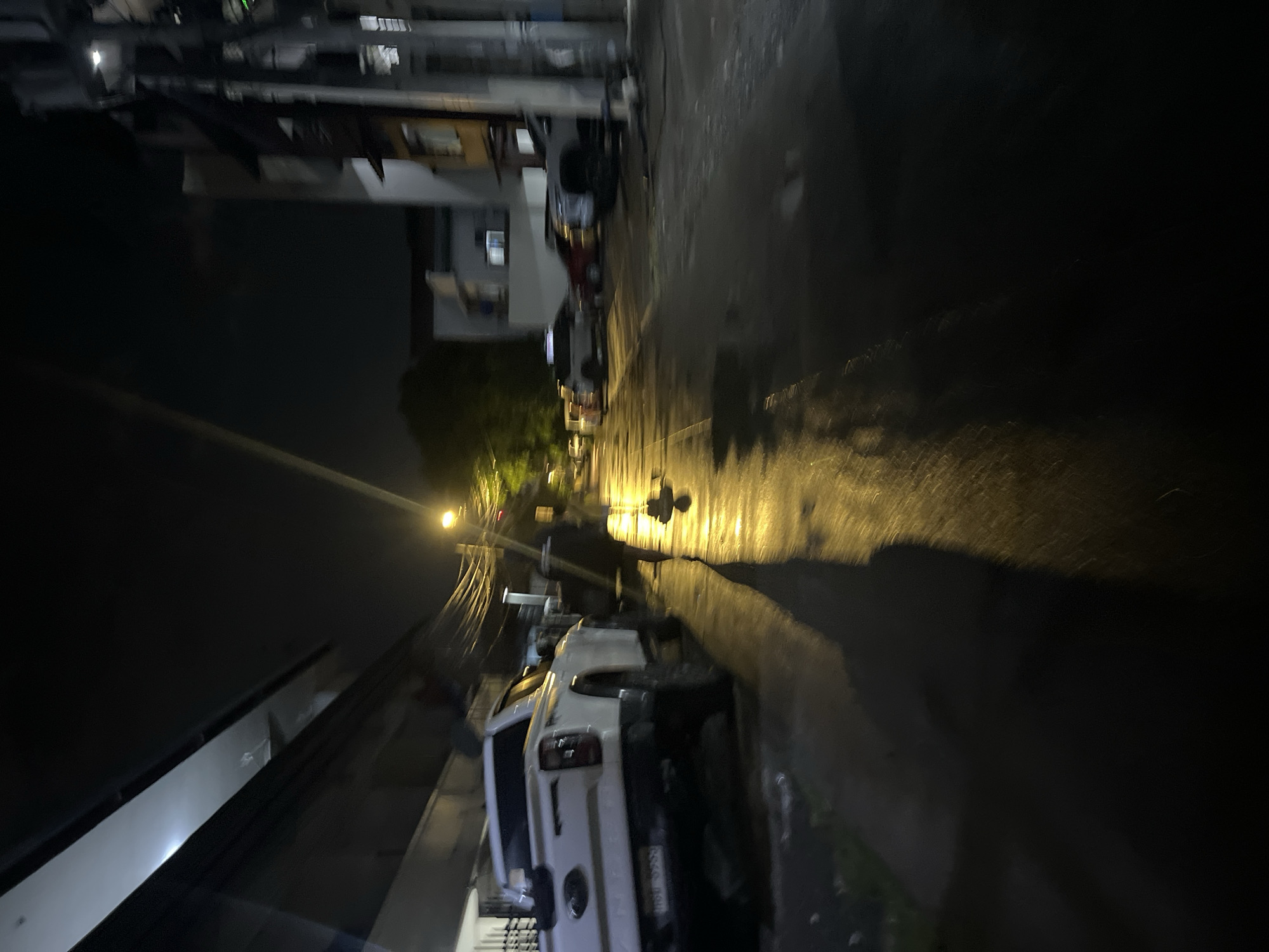 The image shows a street scene at night in Davao City, Philippines. The road surface is wet, reflecting light from a street lamp in the distance. A white pickup truck is parked on the left side of the street. Buildings line both sides of the street, with visible utility poles and wires. A figure is dimly visible walking down the street towards the light. Several other vehicles are parked further down the street.