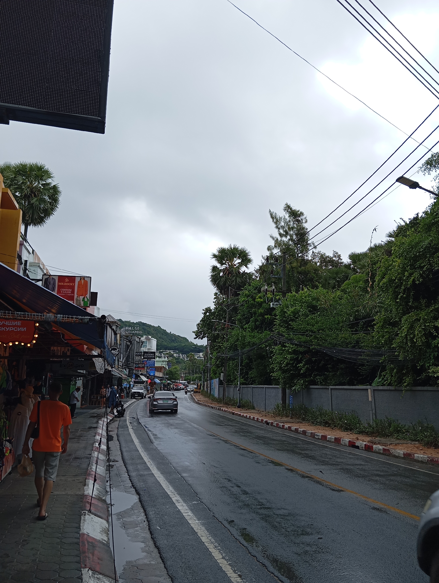 The image depicts a street scene in Karon, Thailand, under an overcast sky, suggesting a cloudy or rainy day. The road is wet, with puddles visible in places, indicating recent precipitation. On the left side of the street, a sidewalk is lined with shops and market stalls, many of which have awnings extending over the pedestrian area. One stall displays clothing, and signs for businesses like "MR. ROCKY CUSTOM TAILOR" and "PALM SQUARE" are visible. A person wearing an orange t-shirt and grey shorts is walking away from the camera on the sidewalk, carrying a plastic bag. Further down the street, several cars are driving on the wet asphalt. The right side of the road is bordered by a grey concrete wall and lush green trees and foliage, with power lines and a lamppost overhead. In the background, a green hill rises, dotted with buildings. The overall impression is of a commercial street in a tropical urban environment.