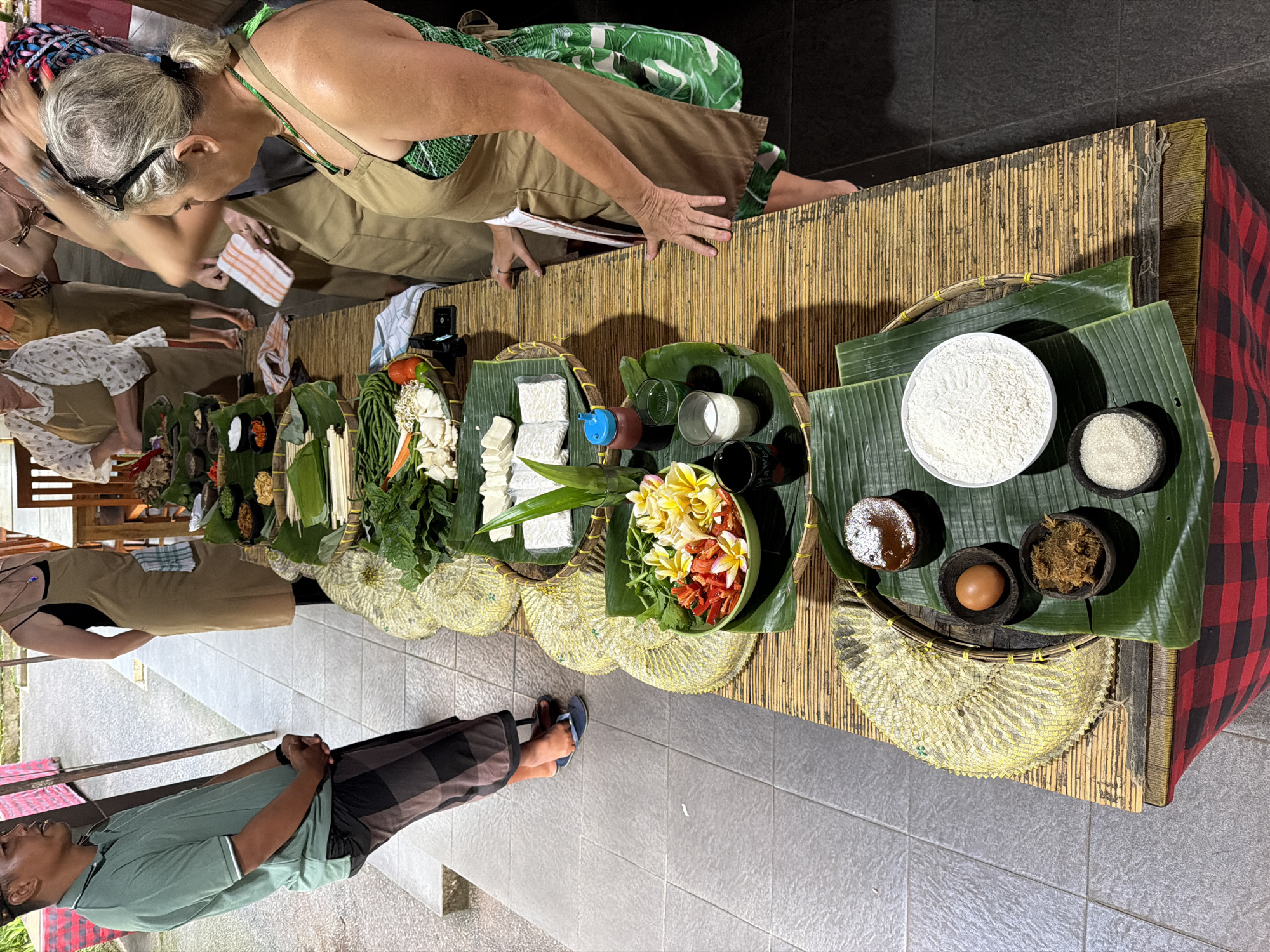 In Ubud District, Indonesia, a culinary class is underway as a man in a green polo shirt and patterned sarong stands to the left, facing a table laden with ingredients. Women in aprons are gathered around the table, preparing for the class. The table is covered with a bamboo mat and a red and black checkered cloth. Various bowls and baskets display fresh produce like long beans, tomatoes, bean sprouts, tofu, and an assortment of colorful flowers. Ingredients such as flour, sugar, spices, and an egg are also laid out. Two women, one with blonde hair and sunglasses on her head, are actively engaged at the table, with one holding a striped towel and the other reaching for an item. The setting appears to be an outdoor or semi-outdoor space, with tiled flooring visible.