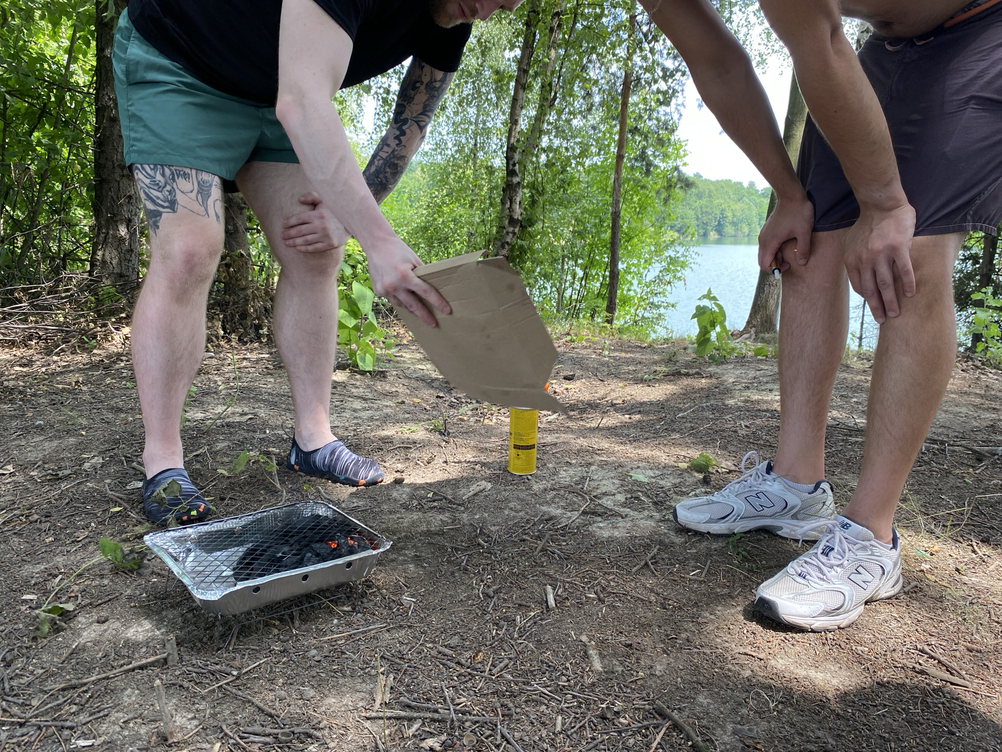 Two individuals are preparing to grill outdoors near a lake in Kaniów, Poland. The ground is covered with dirt, leaves, and small branches. In the foreground, a disposable aluminum grill is filled with burning charcoal. One person, wearing green shorts and tattoos on their left leg, is holding a brown paper bag over the grill. The other person, wearing dark shorts, is standing nearby. A yellow spray can is visible on the ground between them. The background features trees and a body of water, suggesting a natural, possibly forested, recreational area.