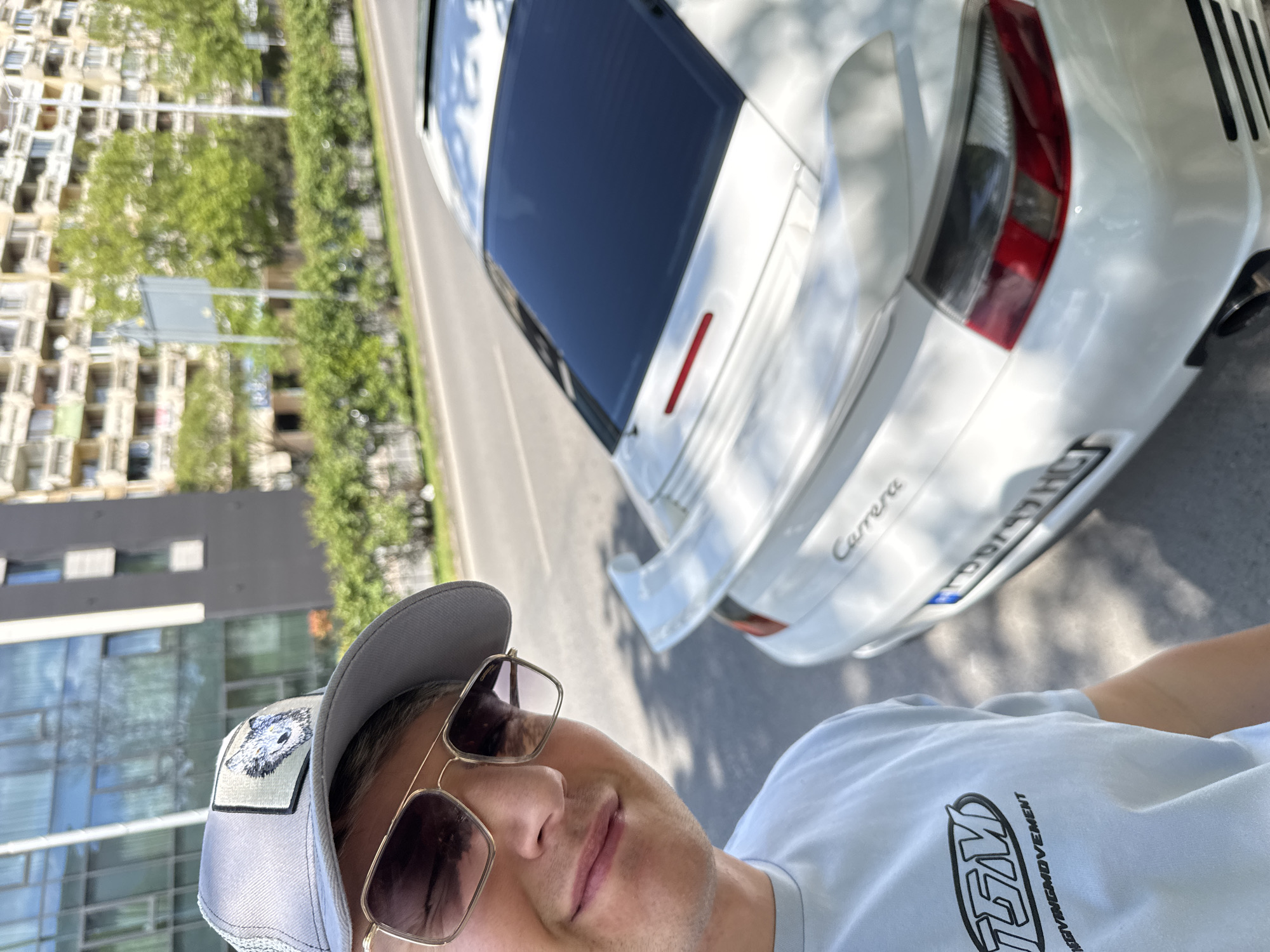 The image is a selfie of a man wearing a gray baseball cap, gold-rimmed sunglasses, and a light blue t-shirt. The background features a white Porsche Carrera parked on a paved road. In the distance, there are buildings and trees, suggesting an urban environment. The license plate of the car is visible. The scene is in Plovdiv, Bulgaria.