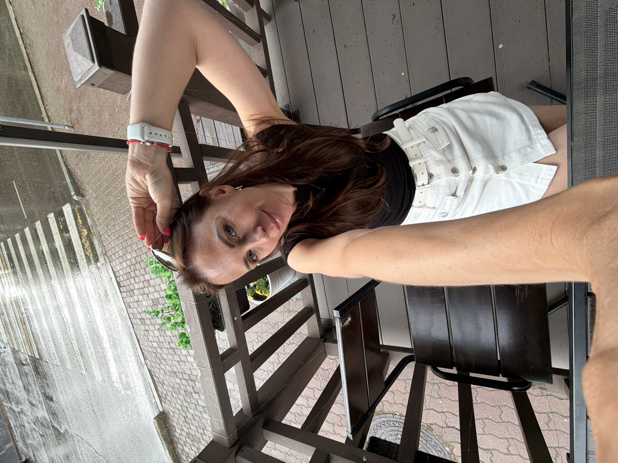 A woman with long brown hair is taking a selfie on a wooden balcony. She is wearing a black tank top and a white denim skirt with a white belt. She has a watch on her left wrist and is holding sunglasses in her right hand. A wooden railing and a part of a chair are visible in the foreground. In the background, there is a crosswalk and pavement. The woman appears to be in an outdoor setting, possibly a cafe or a residential balcony, in the city of Birobidzhan, Russia.