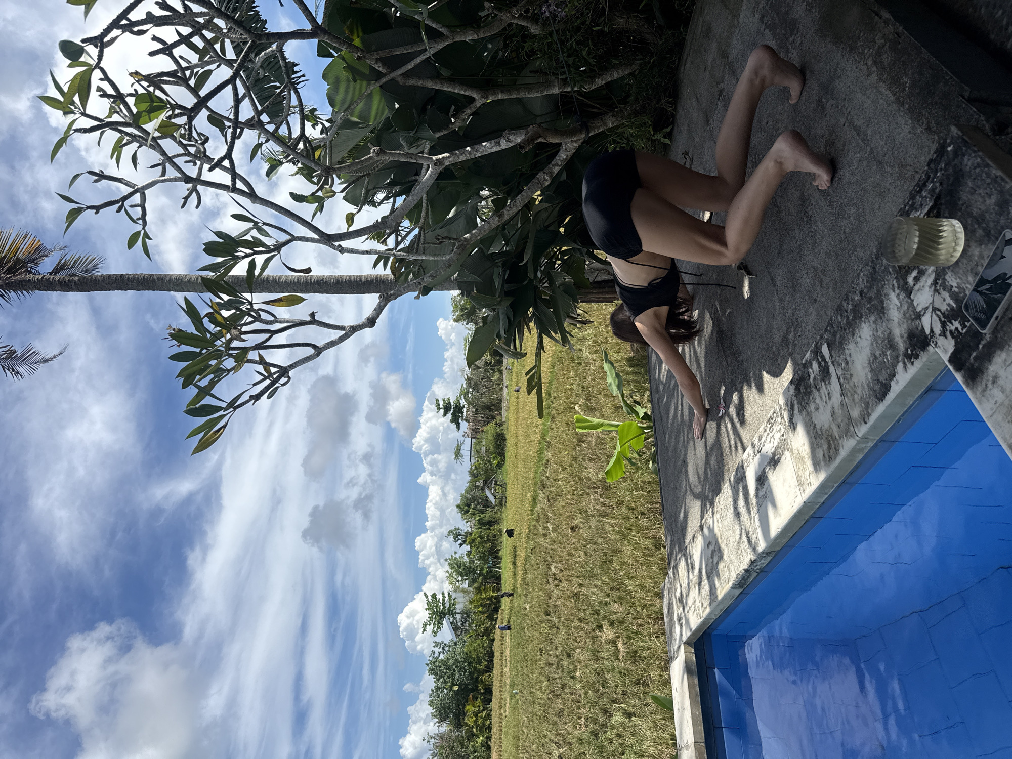A woman in a black bikini is performing a handstand near a swimming pool in Mengwi, Indonesia. The area is lush with trees and foliage. Beyond the pool and the woman, there is a view of a field and a cloudy sky. A glass and a phone are located on the concrete surface next to the woman.