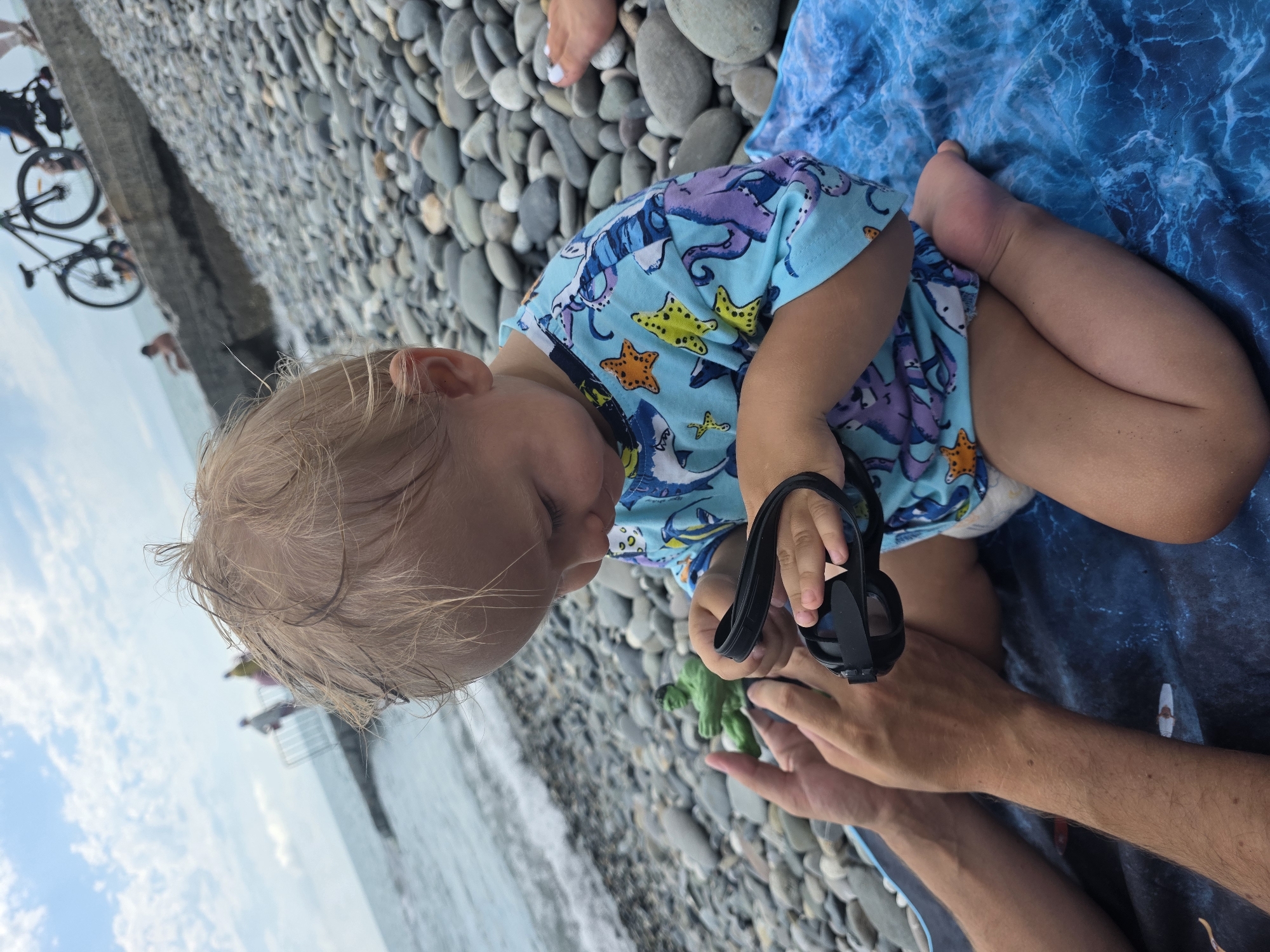 This media file features a young child with blonde hair, wearing a light blue bodysuit adorned with a pattern of sea creatures like octopuses, starfish, and sharks. The child is sitting on a beach covered in smooth, grey pebbles, next to a blue towel with a wave-like pattern. The child is holding a pair of black swimming goggles and a small green toy. An adult's hands are visible, interacting with the child and the toy.
In the background, the sea is visible with gentle waves lapping at the shore. Further back, a metal railing suggests a pier or boardwalk. On the left side of the image, two bicycles are parked near a concrete structure, and a person is standing in the water. The sky is partly cloudy with a bright, diffused light, indicating it is daytime, likely late morning or afternoon. The overall atmosphere seems relaxed and summery. The context suggests a day at the beach in Sochi, Russia. No visible text is present in the image.
