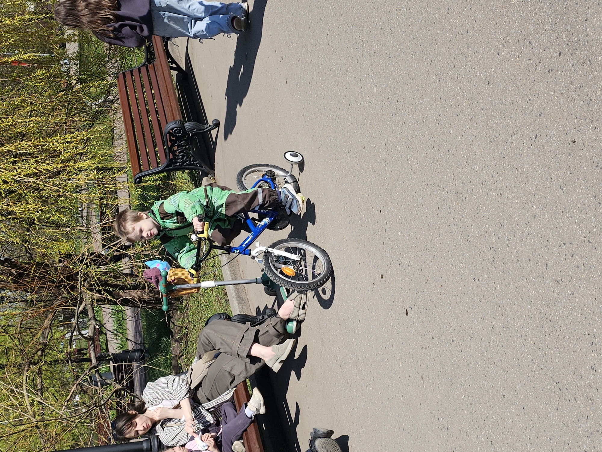 A medium-shot photo captures a scene in what appears to be a park, potentially in the city of Moscow, Russia. A young boy wearing a green jacket is positioned near a blue bicycle, which is lying on its side on the pavement. The child’s attention seems to be directed towards the bicycle. Adjacent to the bicycle and the boy, there are two park benches. One bench is occupied by an adult woman who appears to be lying down or resting, while another woman sits next to her, holding a young baby. On another bench, a girl is standing. The environment includes greenery and trees, suggesting a relaxed outdoor setting. The surface is a paved walkway or road.